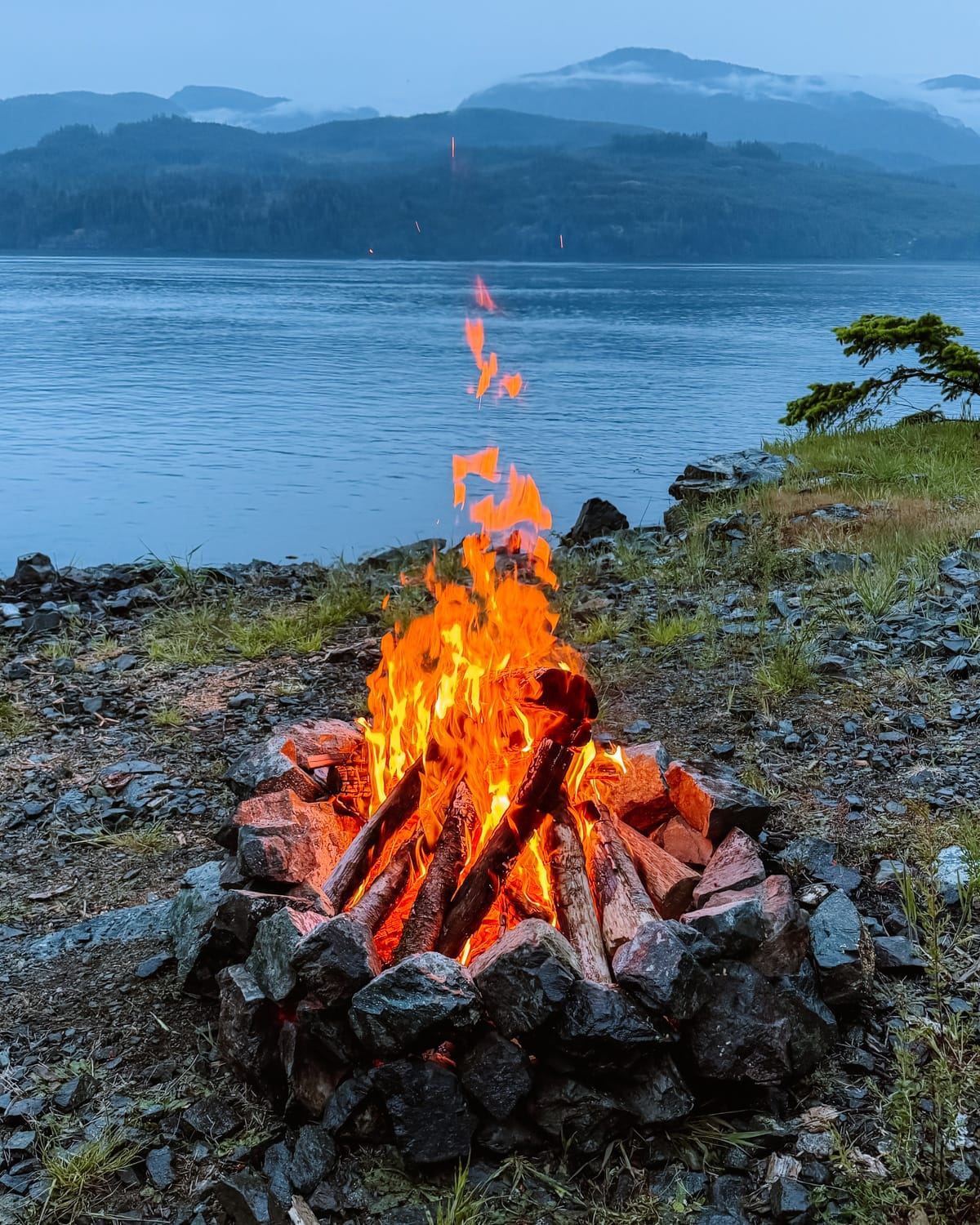 A blazing campfire on a rocky shore lights up the dusk on Quadra Island, with calm water and misty mountains in the background.