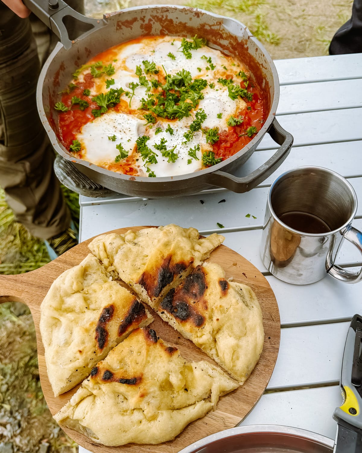 A rustic meal setup outdoors, featuring a skillet of shakshuka topped with herbs and a wooden board with warm, fluffy flatbread.
