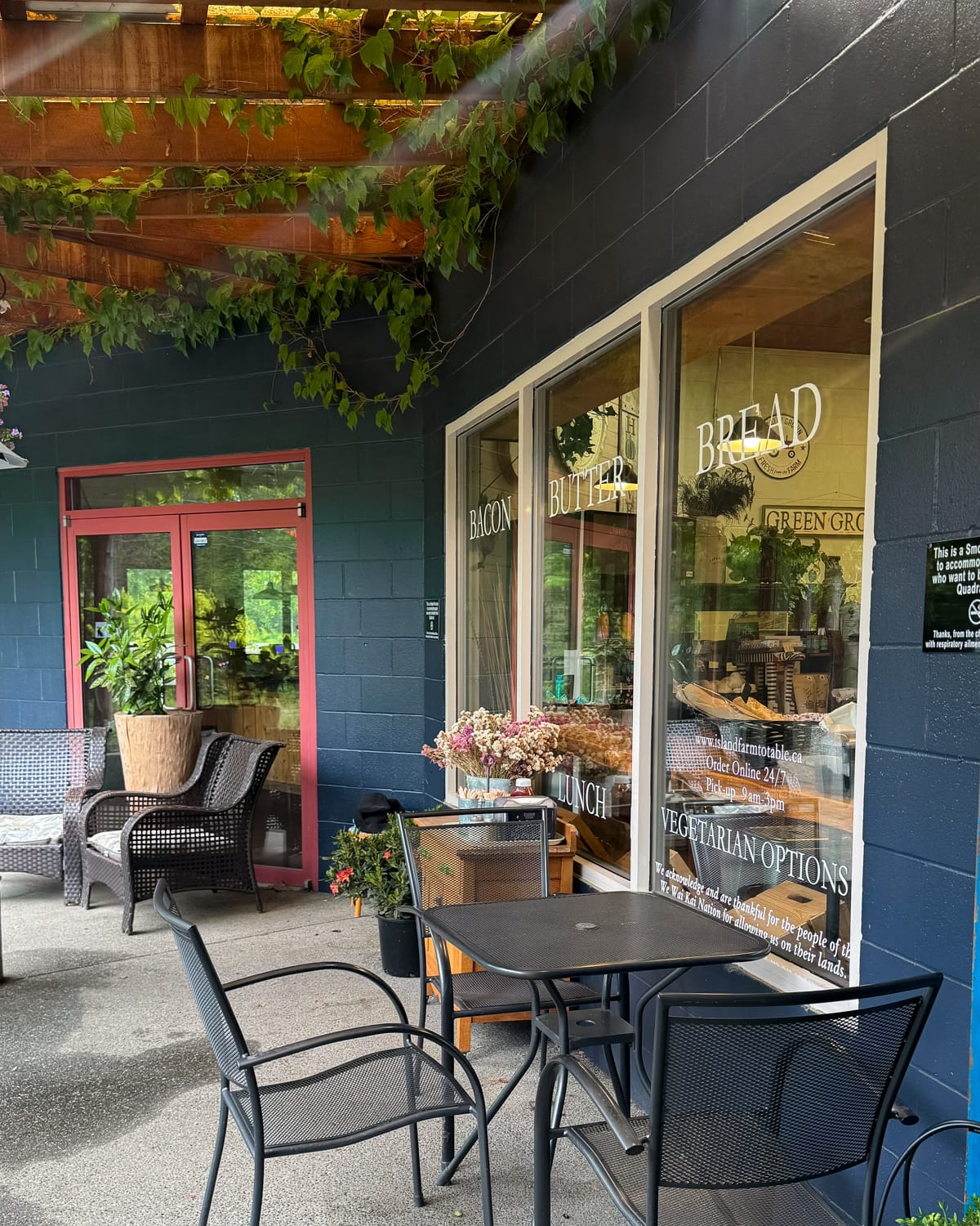 Outdoor patio of Island Farm and Table cafe on Quadra Island with ivy-covered beams, metal furniture, and large window signage reading “Bacon, Butter, and Bread” and “Vegetarian Options.