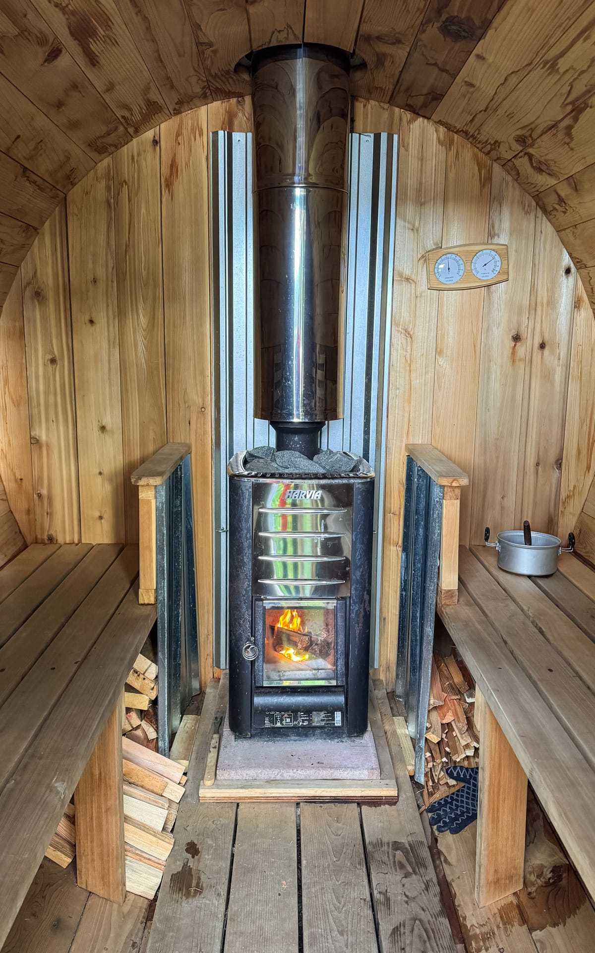 Close-up of a cozy wood-burning stove heating a barrel sauna, with wooden benches and stacked firewood on both sides at Embrace Sauna on Quadra Island.
