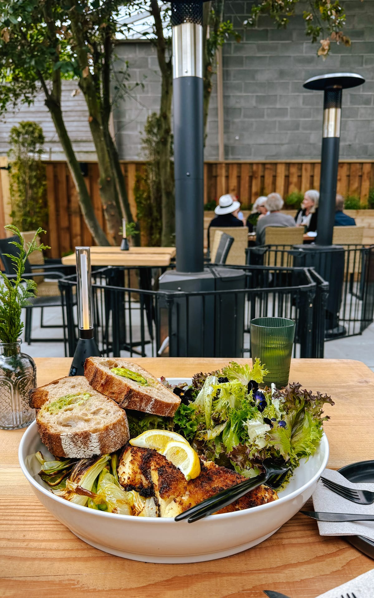 Grilled fish with lemon slices, fresh greens, and toasted bread served on a patio table, with diners in the background at Feast on Salt Spring.
