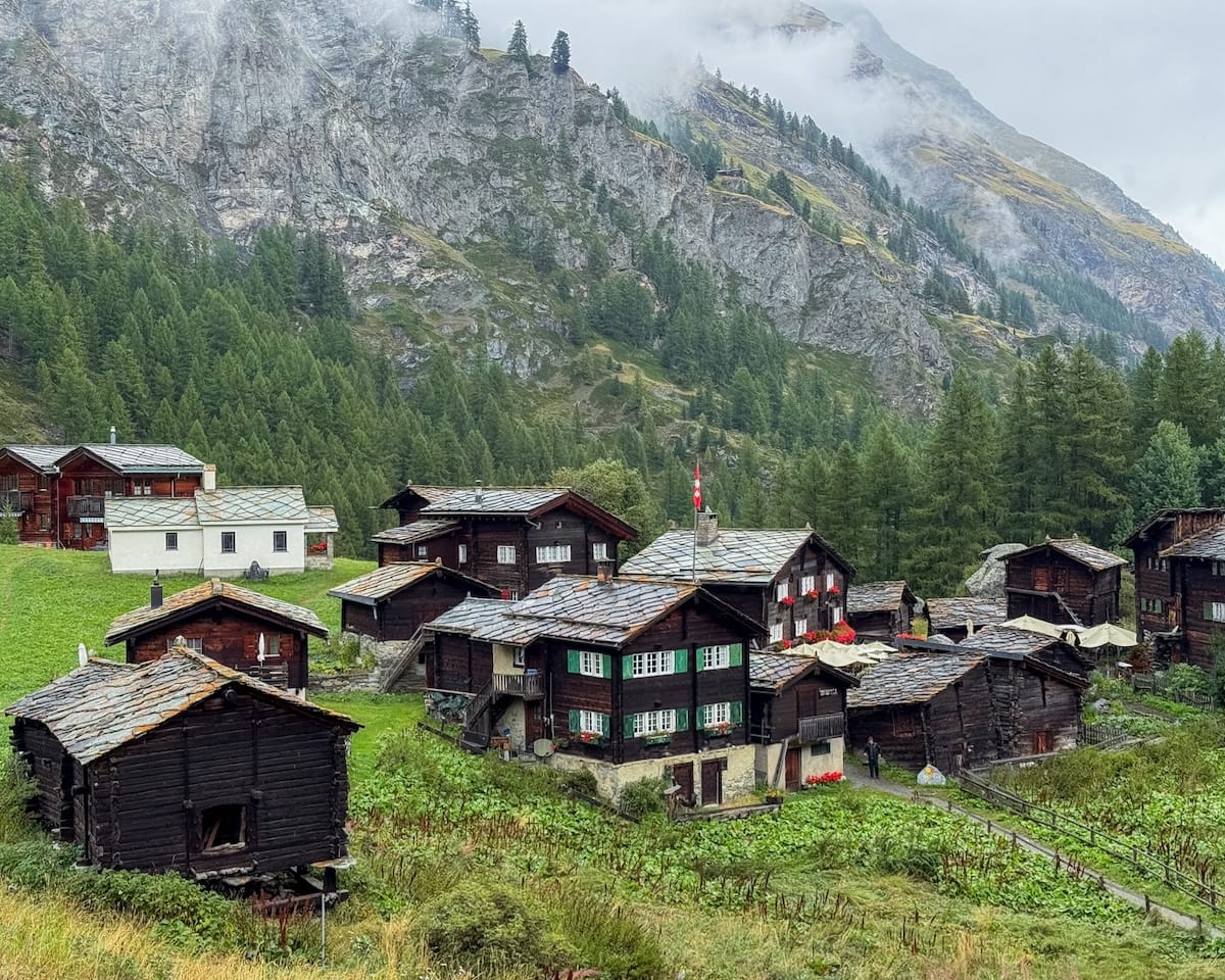 Rustic Zum See village with traditional wooden chalets nestled in a misty valley, surrounded by towering cliffs and greenery.