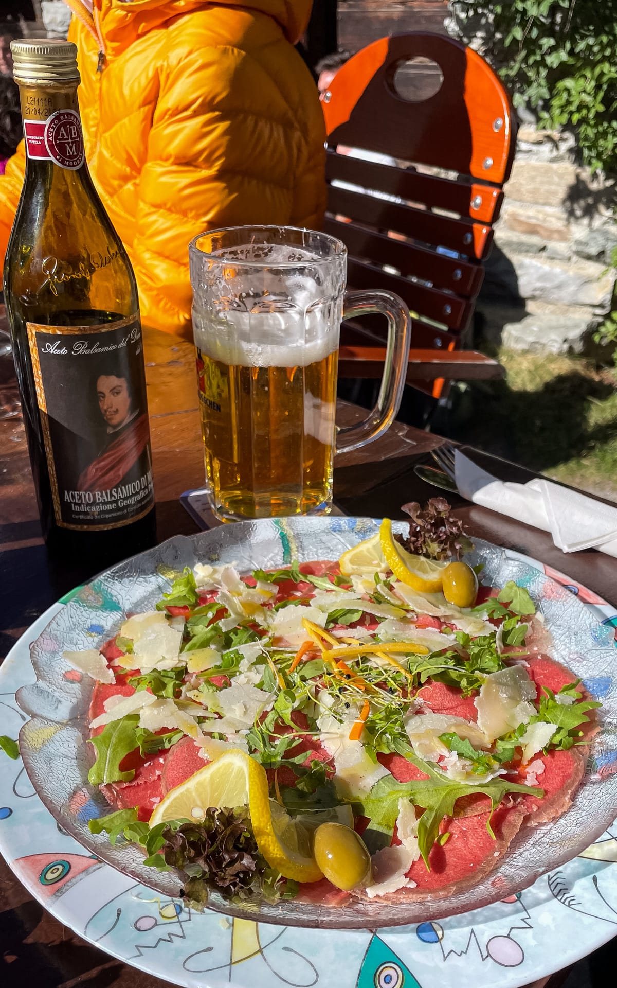 Beef carpaccio garnished with arugula and parmesan, served with balsamic vinegar and beer on a rustic outdoor patio.