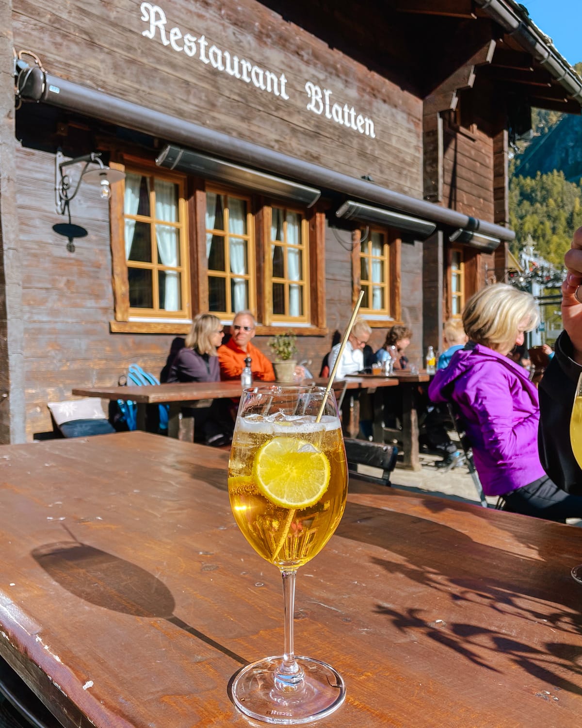 A refreshing spritz with lemon at an outdoor table beside Restaurant Blatten in a wooden chalet.