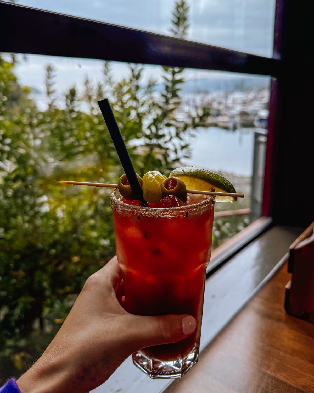 Classic Caesar cocktail garnished with olives and pickles, held up against Moby's harbour view backdrop.