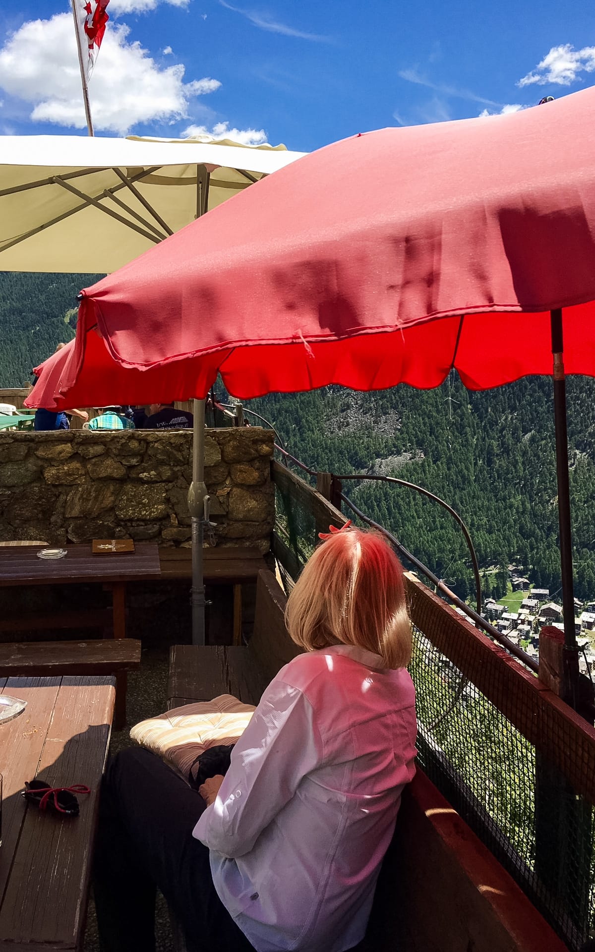 Woman enjoying outdoor dining under a red umbrella on a sunny terrace, overlooking Zermatt below.