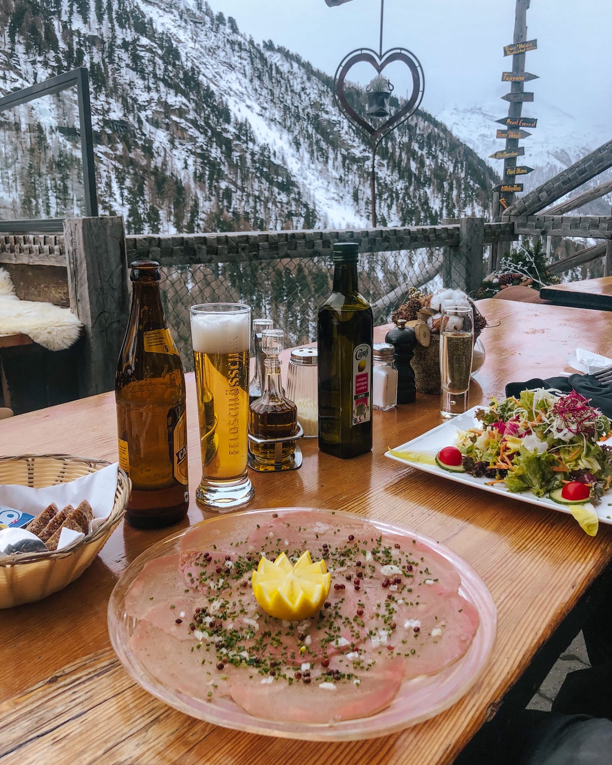 Outdoor mountain dining at Findlerhof with cured meat, fresh salad, and beer overlooking snow-covered cliffs..