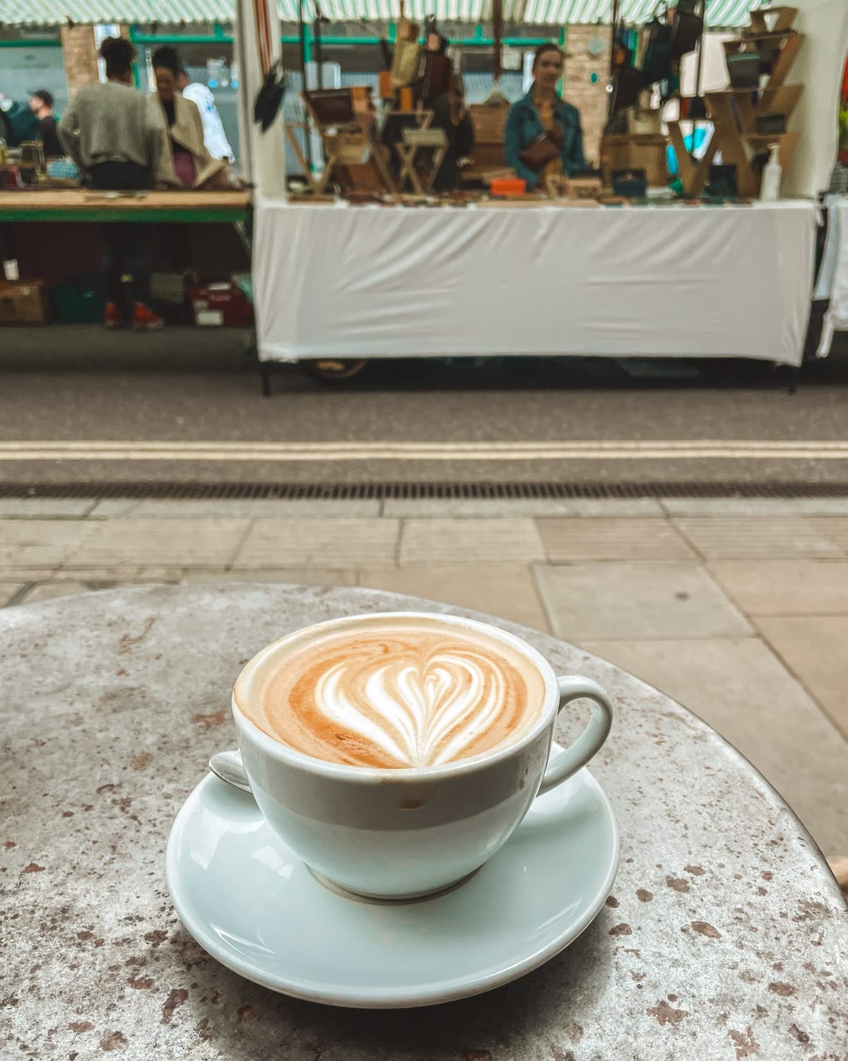 A cappuccino with heart-shaped foam art sits on a cafe table at Broadway Market, with stalls in the background.