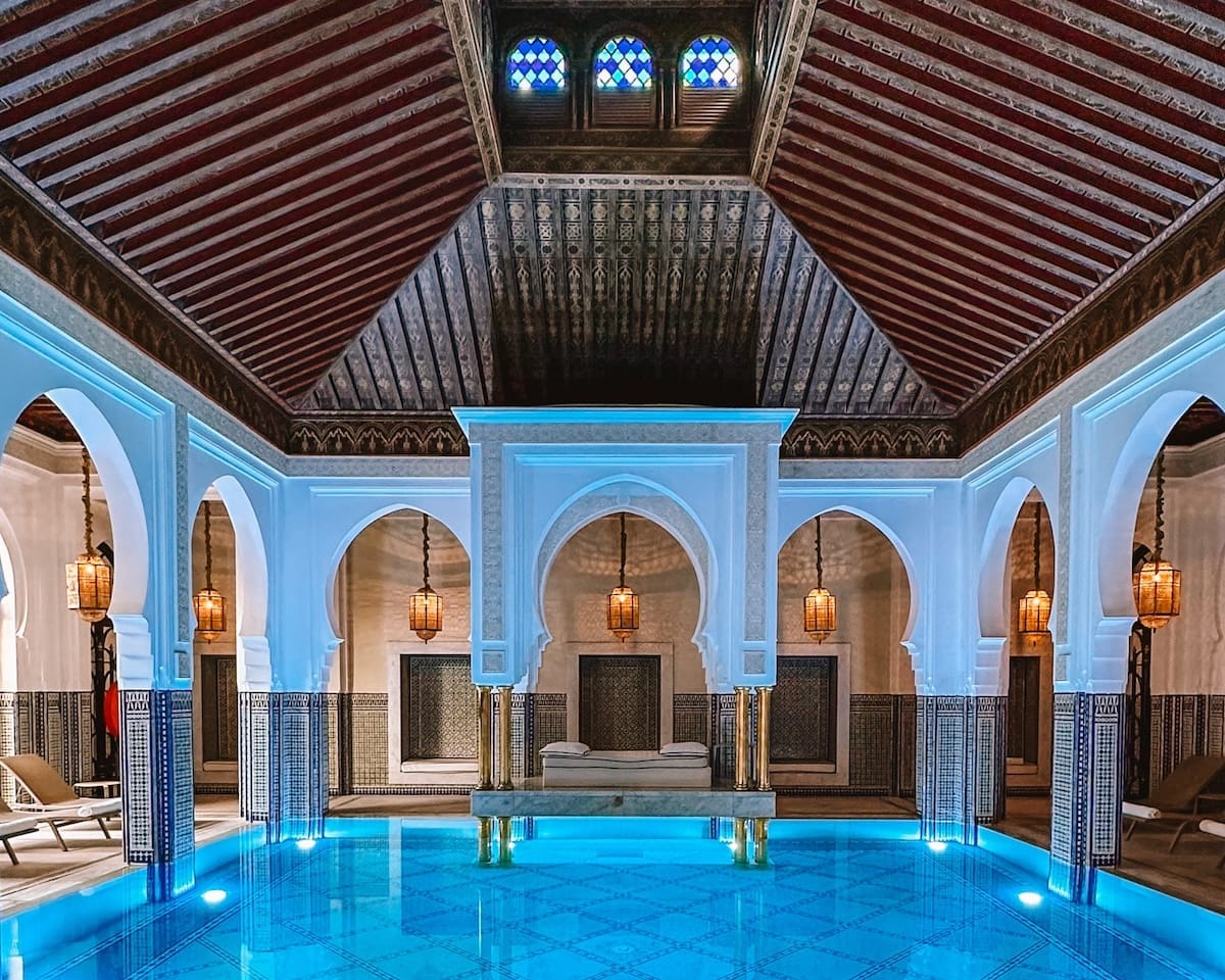 Elegant indoor pool with arched white columns, intricate tilework, and warm lantern lightin at La Mamounia in Marrakech, creating a serene spa-like atmosphere.