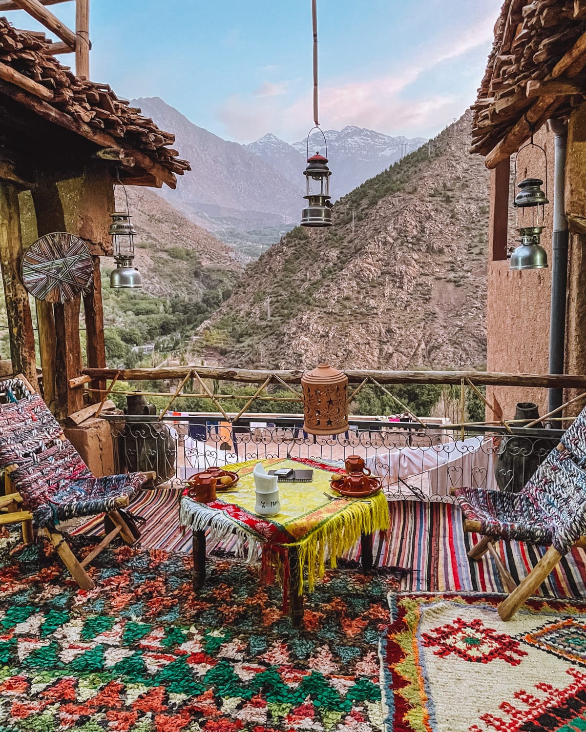 Cozy guesthouse balcony with colourful Berber rugs, low chairs and a tea setup, overlooking the Atlas Mountains at sunset.