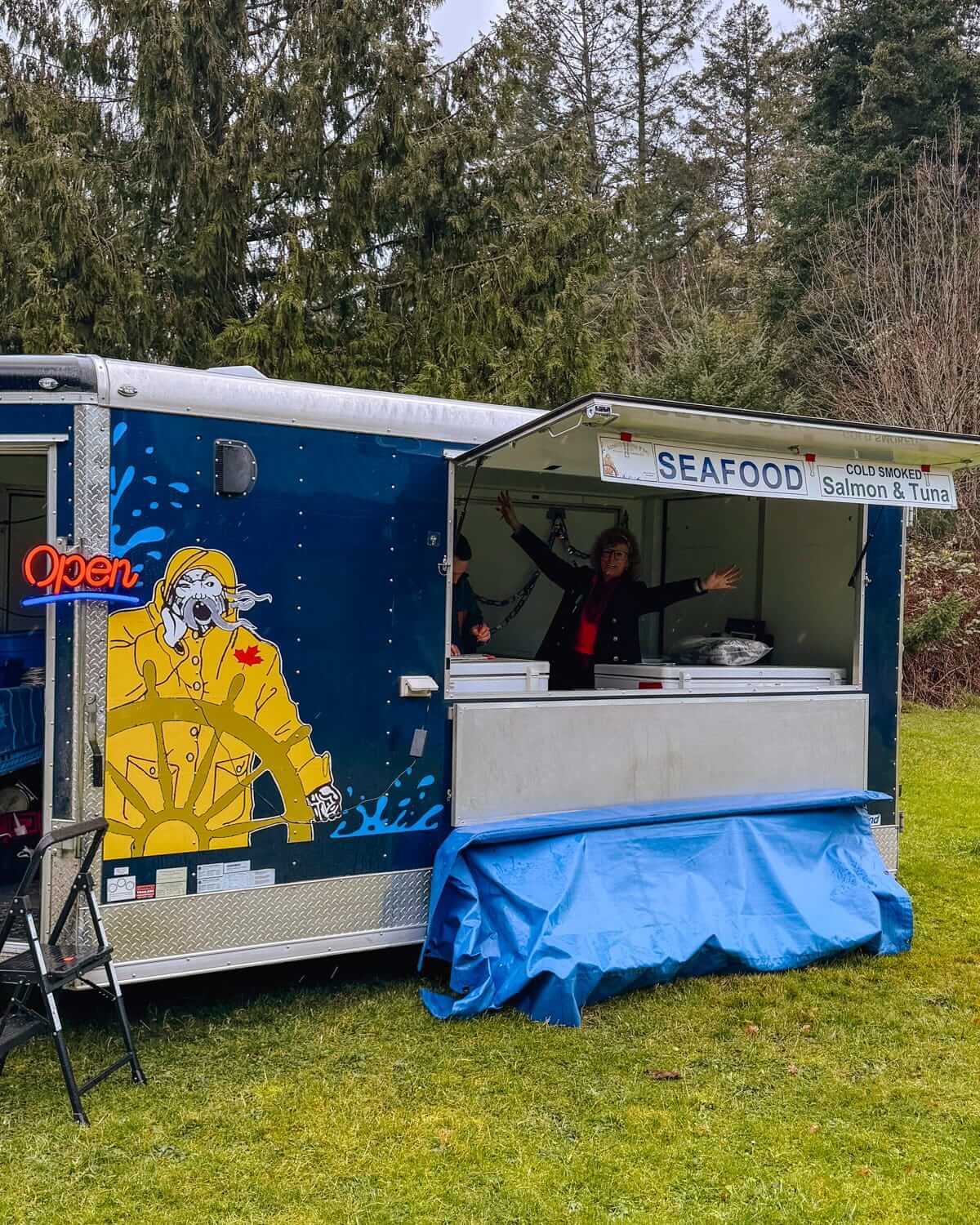 Seafood vendor in a vibrant blue food truck with a fisherman illustration in a forested setting.