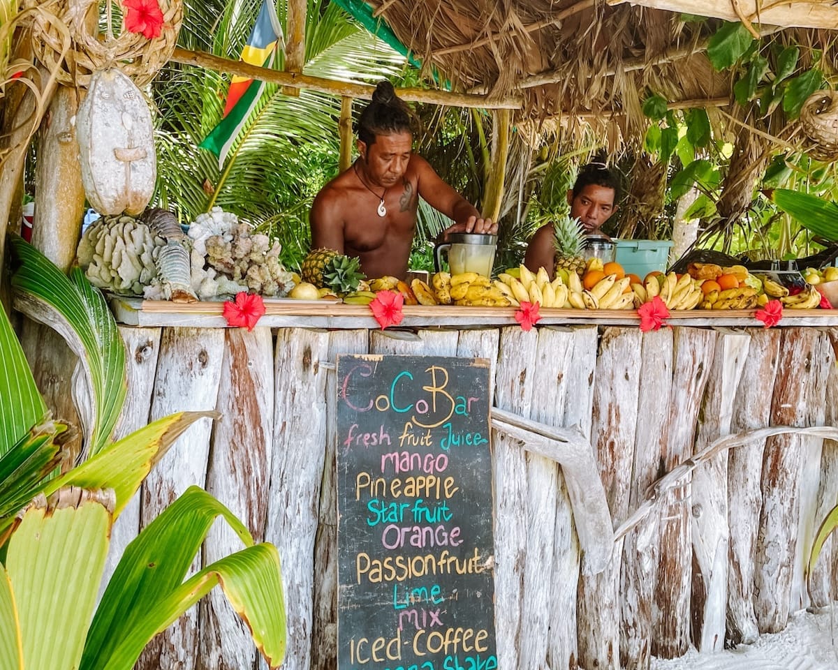 Beachside fruit stand in Seychelles with fresh mangoes, pineapples, and passion fruit, run by locals under a thatched roof with tropical decor/