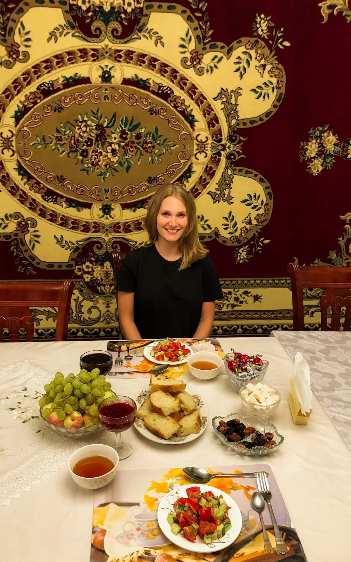 Cec enjoying a Kyrgyz dinner with bread, salad, fruit, and tea, in a traditional, richly decorated dining room of a guesthouse.