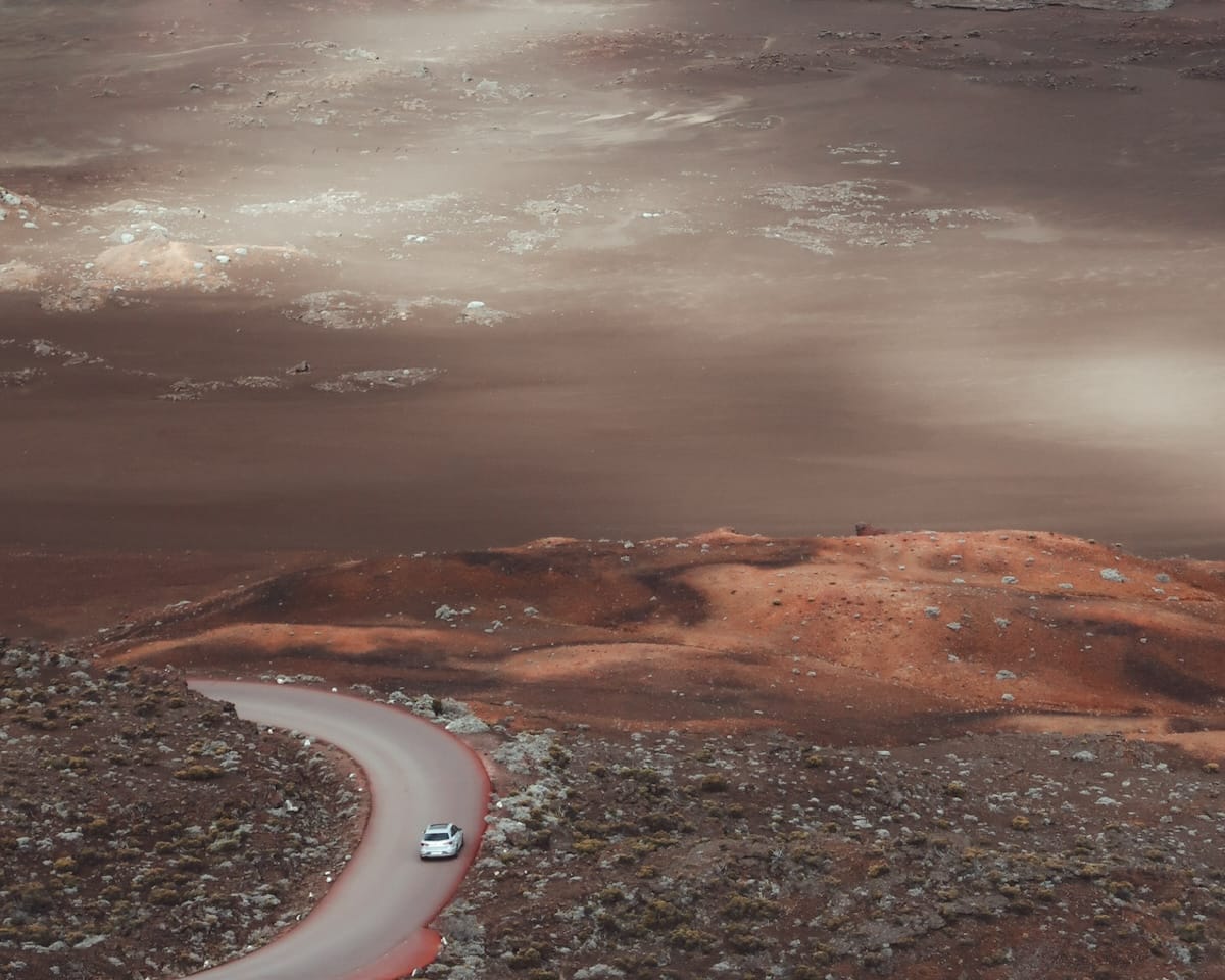 A winding road cuts through a barren, Mars-like volcanic landscape, with a lone white car navigating the rugged terrain on the Route du Volcan.