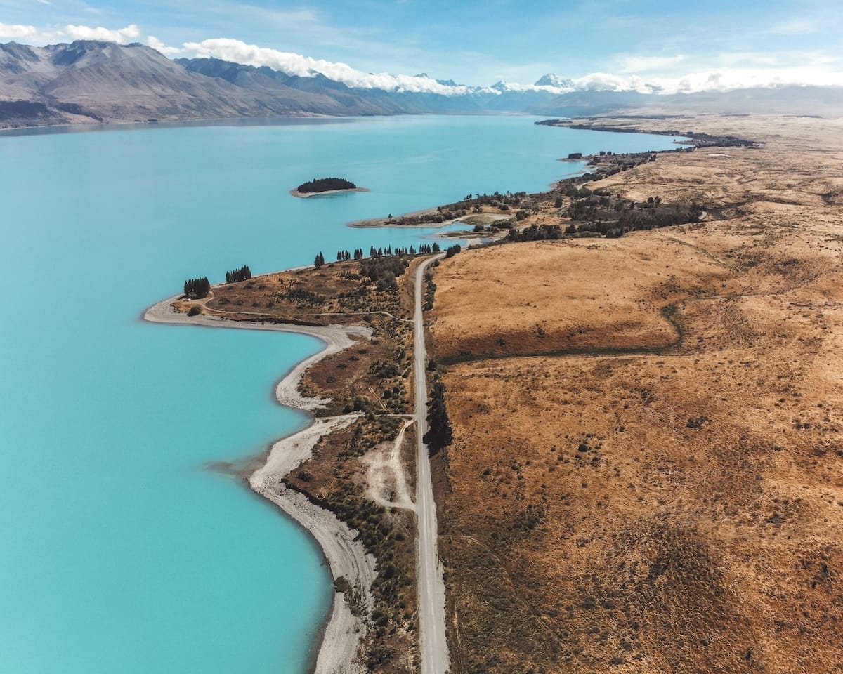 Aerial view of Lake Pukaki on the South Island of New Zealand, with turquoise water, dry terrain, winding road, and snow-capped mountains beyond.