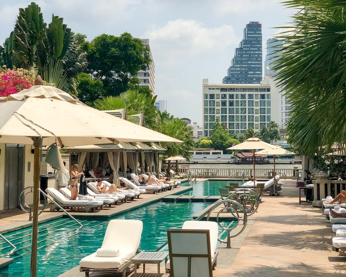 A riverside pool lined with sun loungers and cabanas at The Peninsula, with Bangkok’s modern skyline rising in the background.