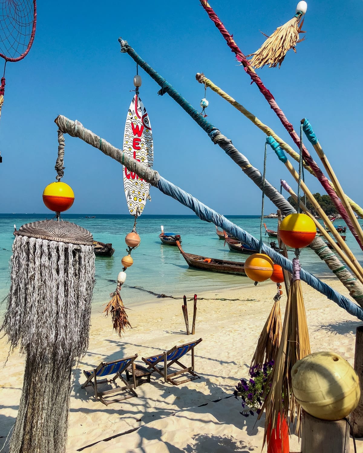 A colourful beachside installation with wrapped poles, buoys, and a "Welcome" surfboard, overlooking turquoise waters on Koh Lipe island..