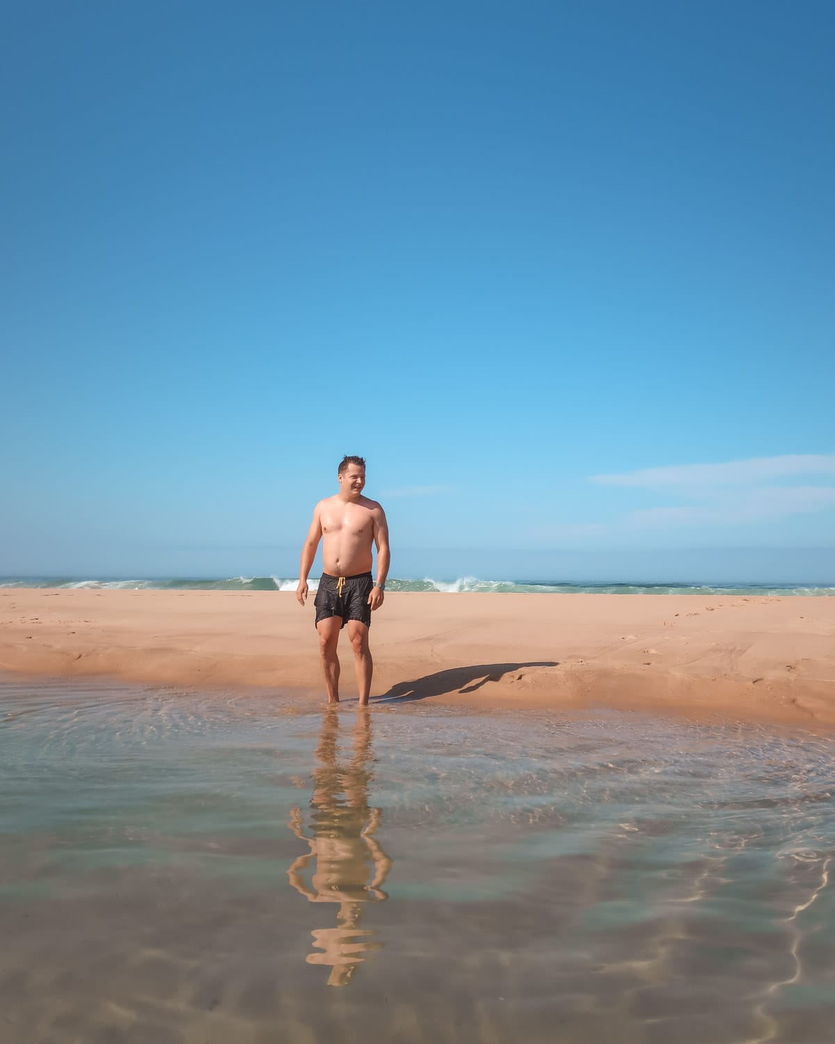 Ari stands in a shallow tidal pool on a sandy beach in Cap Ferret, with ocean waves and a clear blue sky in the background.