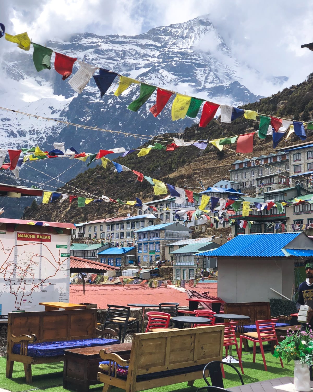 Colorful prayer flags hang above outdoor seating in Namche Bazaar with a snowy mountain backdrop.