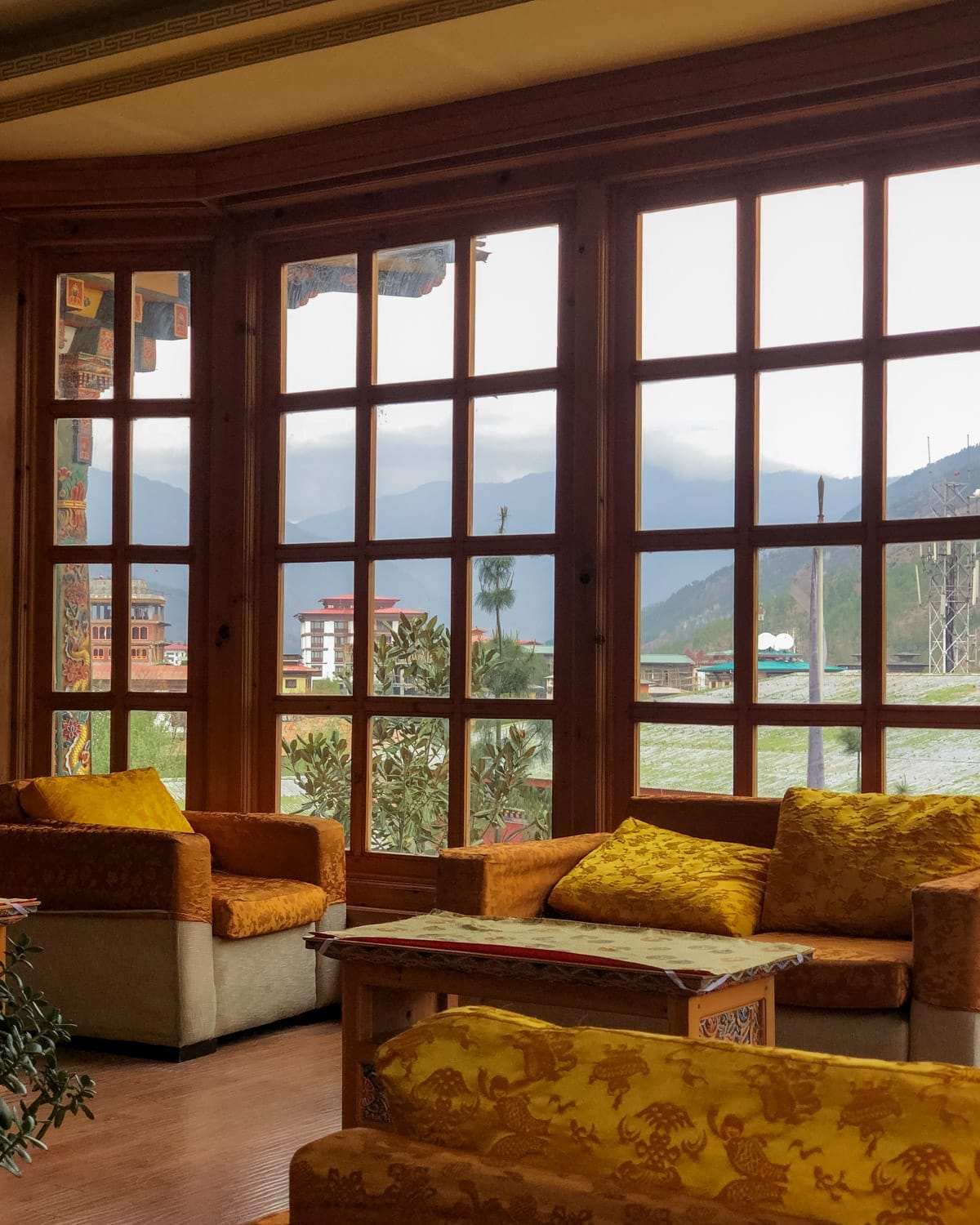 Cozy lounge at Namgay Heritage Hotel with plush mustard-yellow seating, and large wooden windows framing a misty mountain view in the distance.