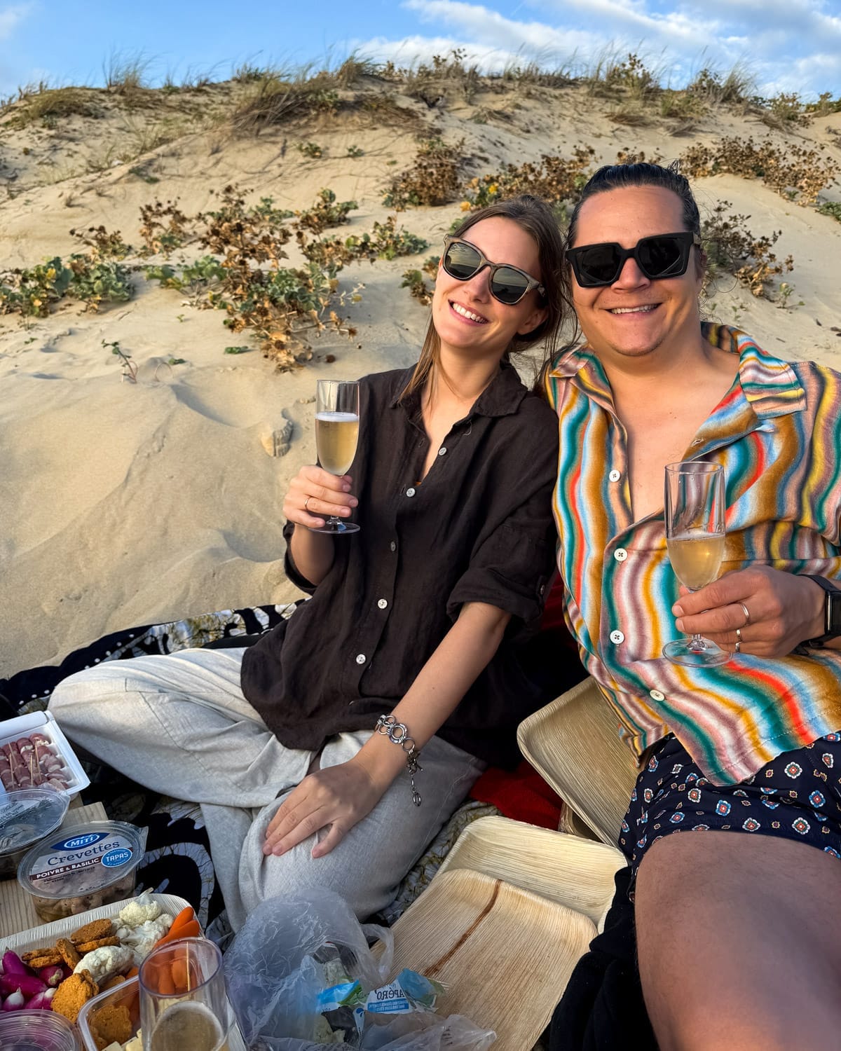 Cec and Ari enjoying a picnic on a sandy dune, sipping champagne and eating picnic bites under the evening sky.