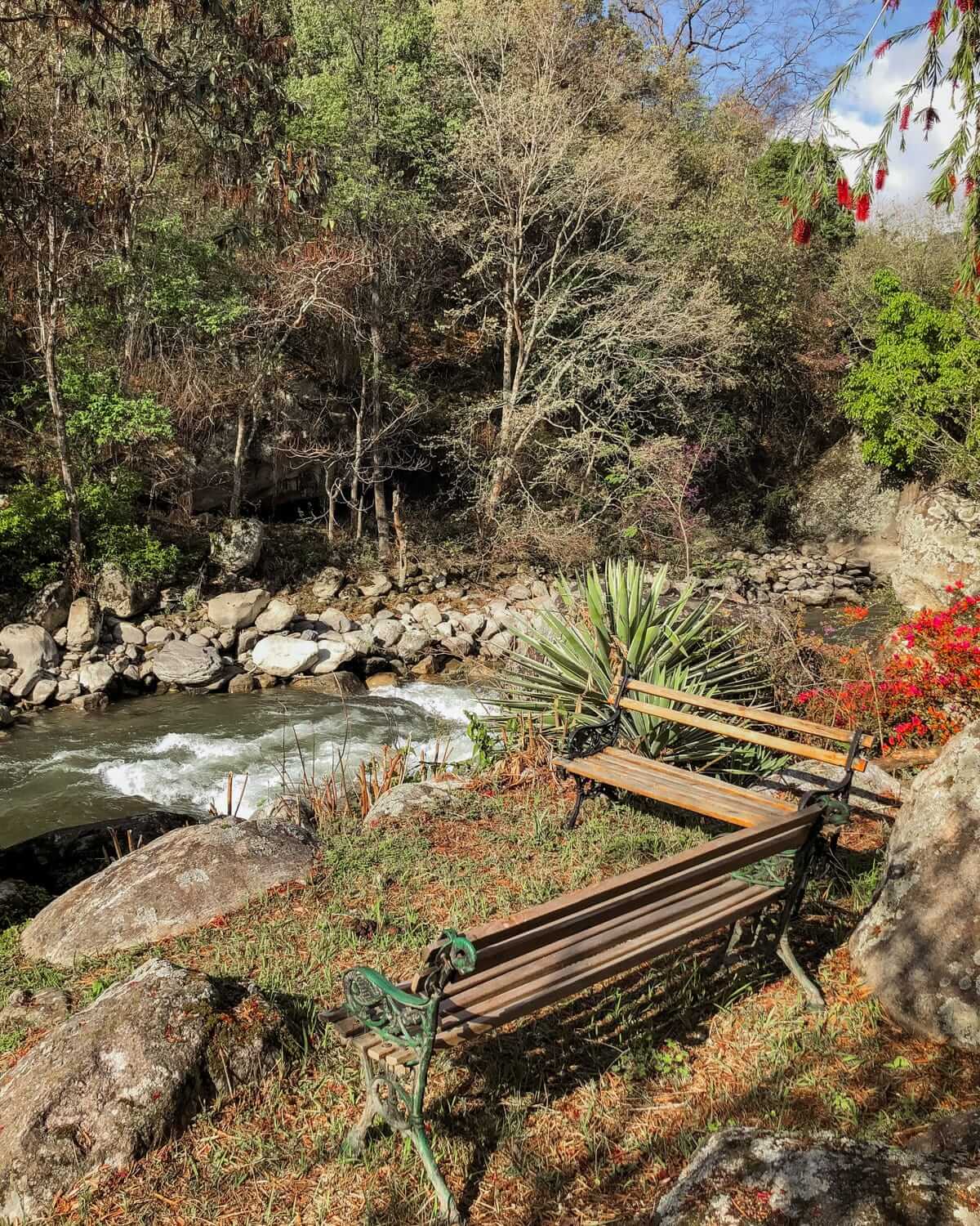 Two rustic wooden benches by a river at Kichu Wangdu Resort, surrounded by lush greenery and red flowers, offering a peaceful nature escape in Bhutan.