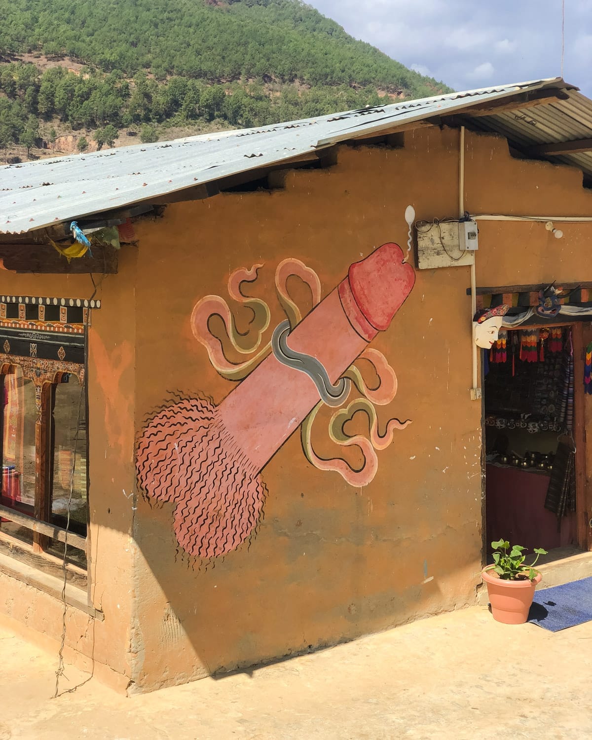 Phallic mural on a Bhutanese house in Punakha, a symbol of fertility and protection, painted on an earthy-toned wall near a souvenir shop.