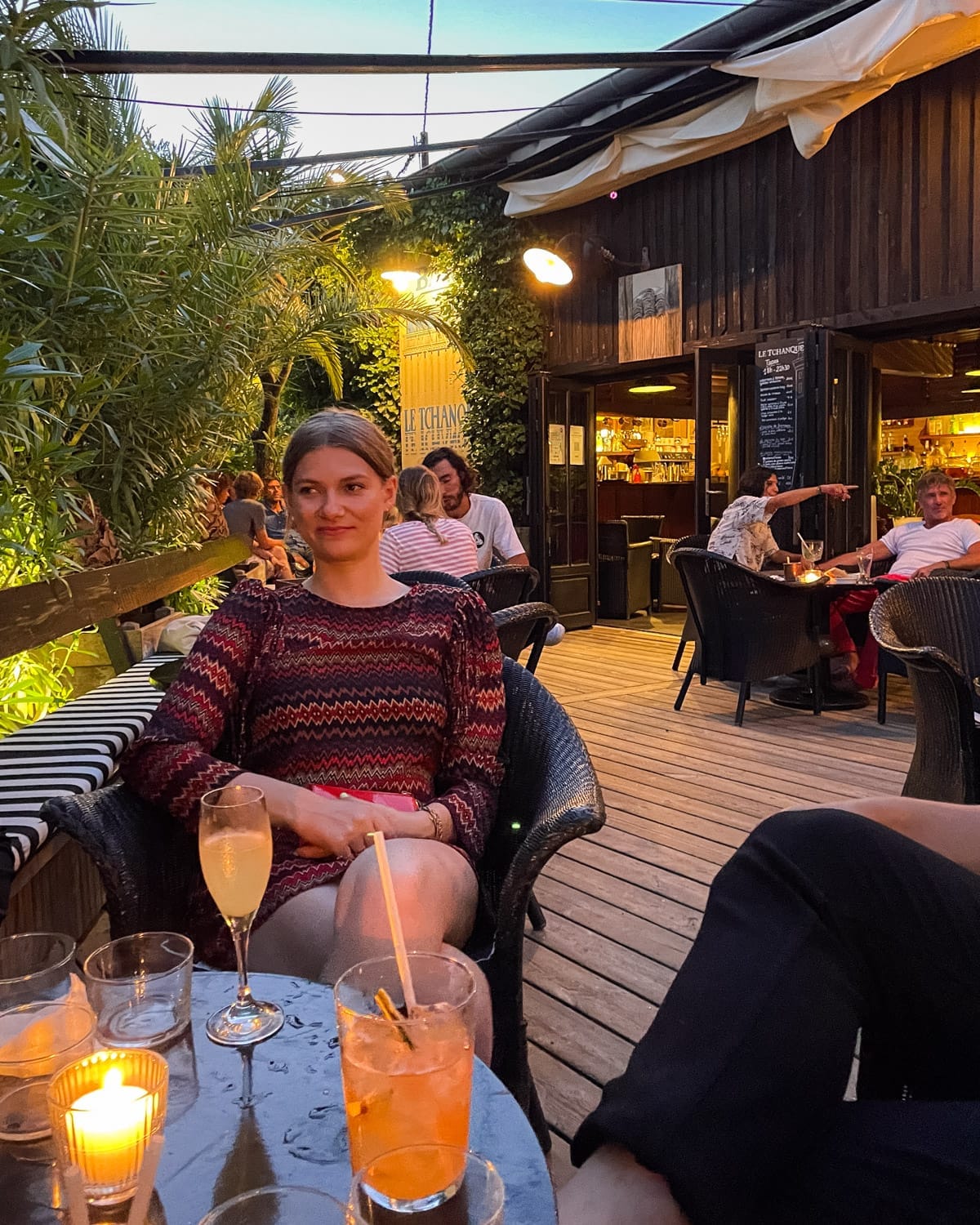 A woman enjoys cocktails outside at Bar Le Tchanque with wooden decking and warm evening lighting.