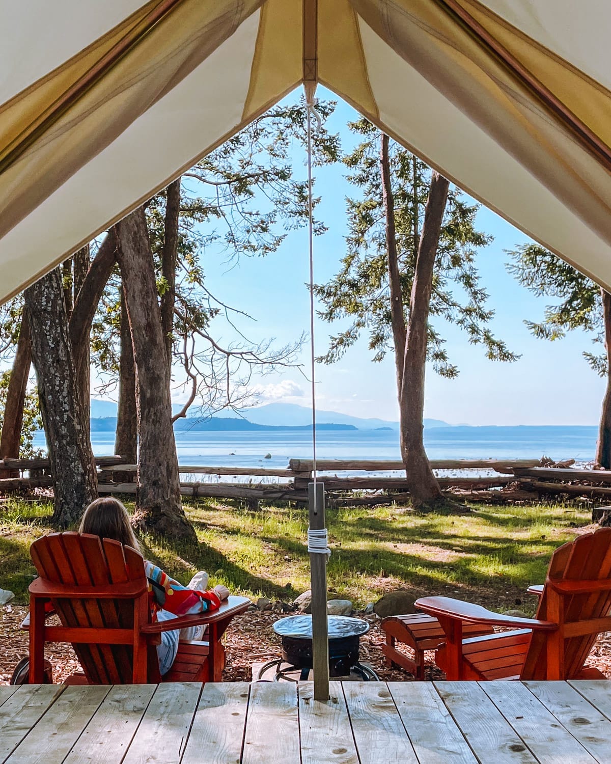 Cec sitting in a red cottage chair under a canvas tent, gazing at a tranquil ocean framed by tall trees and distant mountains on Hornby Island.
