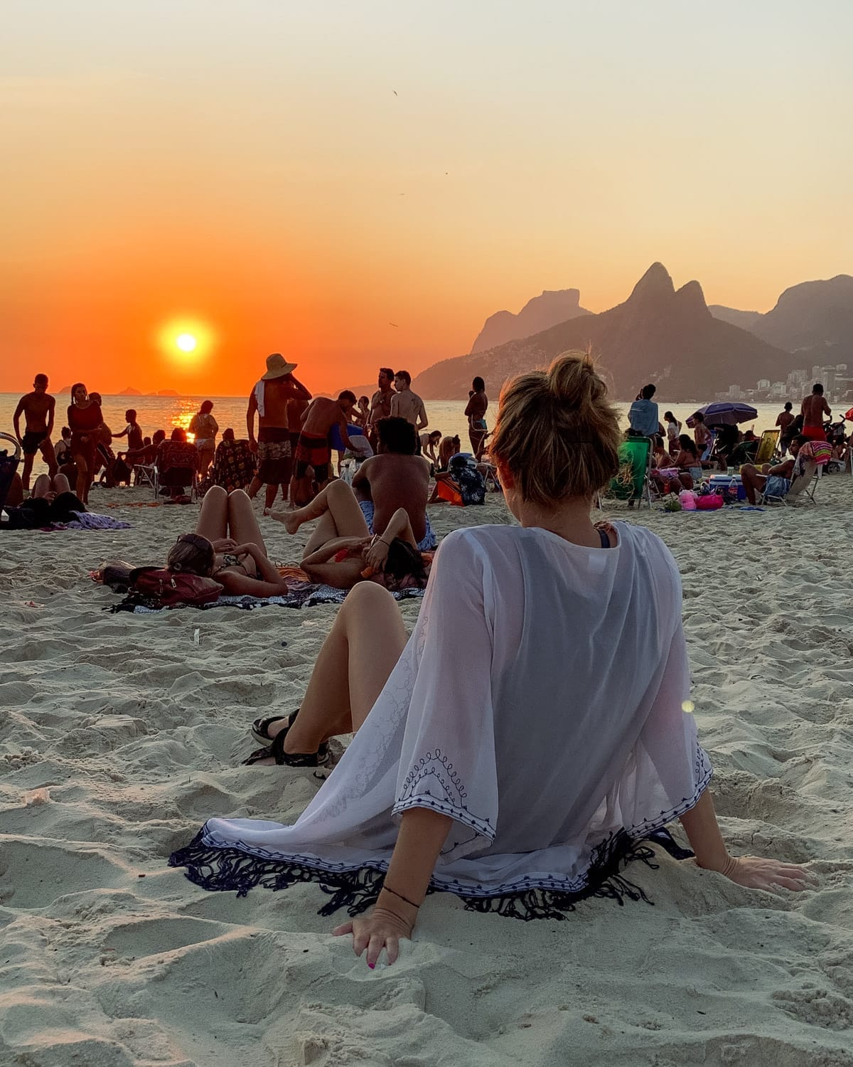 Crowds gather on Ipanema Beach at sunset, as Cec in a beach cover-up relaxes in the sand facing Rio’s golden horizon.