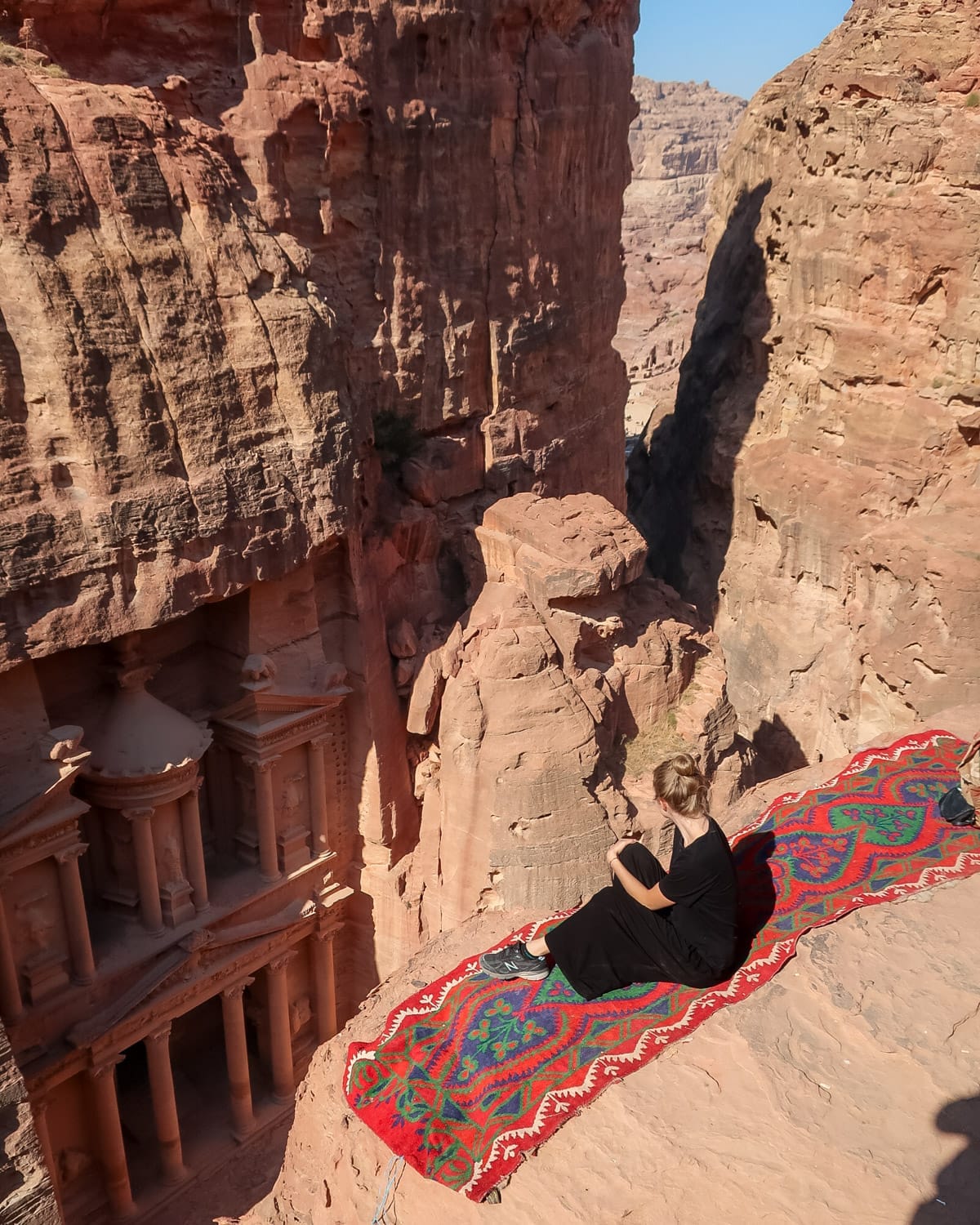 Cec sits on a red-patterned rug at a cliff's edge, overlooking the carved façade of the Treasury at Petra.