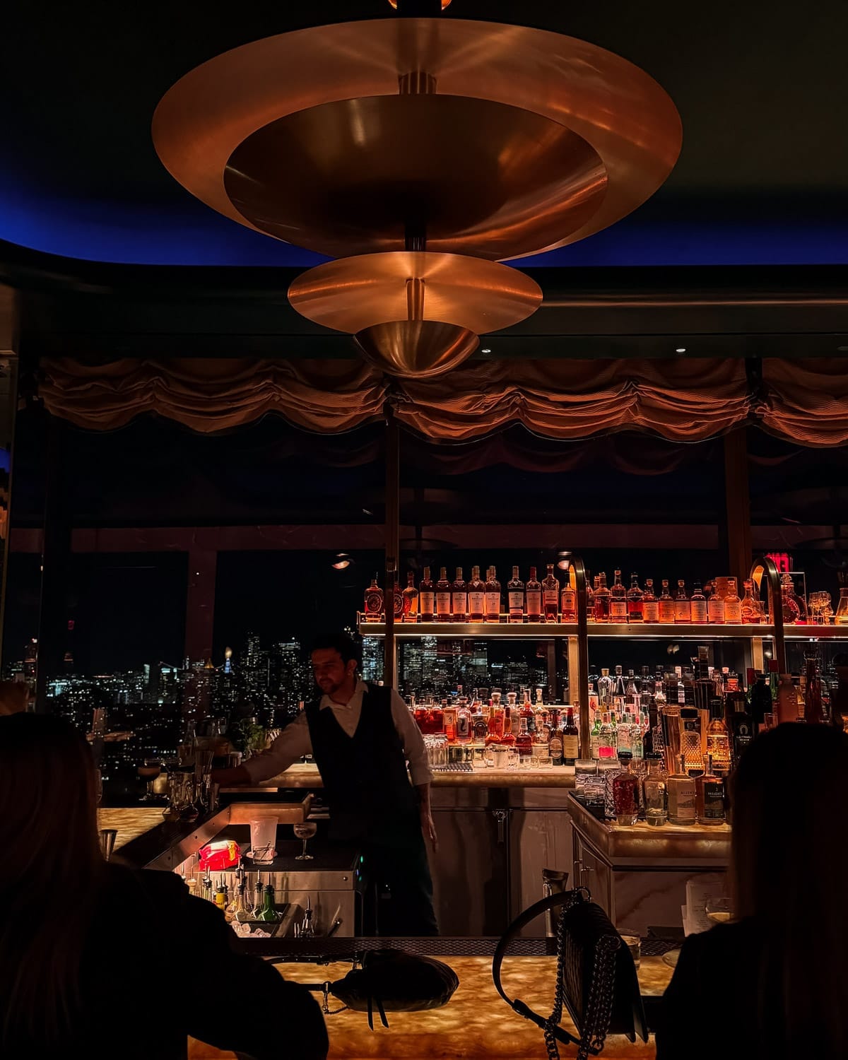 A bartender serves drinks at a dimly lit rooftop bar in Manhattan with a golden chandelier and view of the city skyline at night.