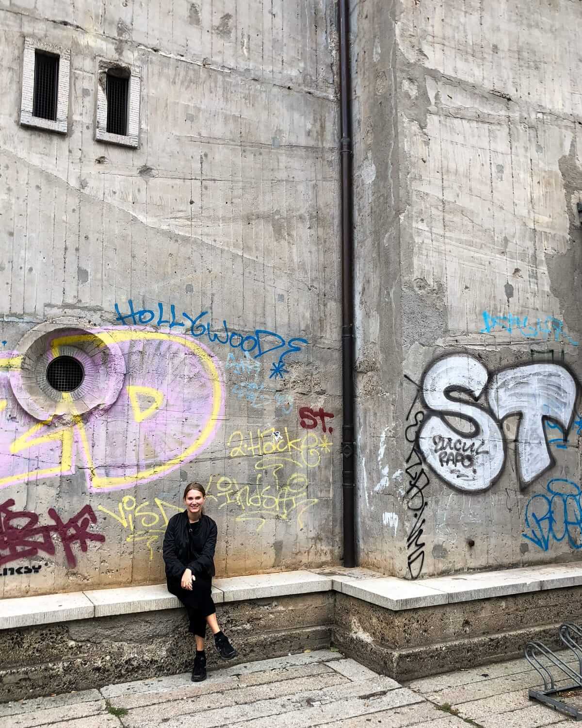 Cec sitting on a bench against a graffiti-covered concrete bunker in Berlin, featuring colourful street art and rough textures.
