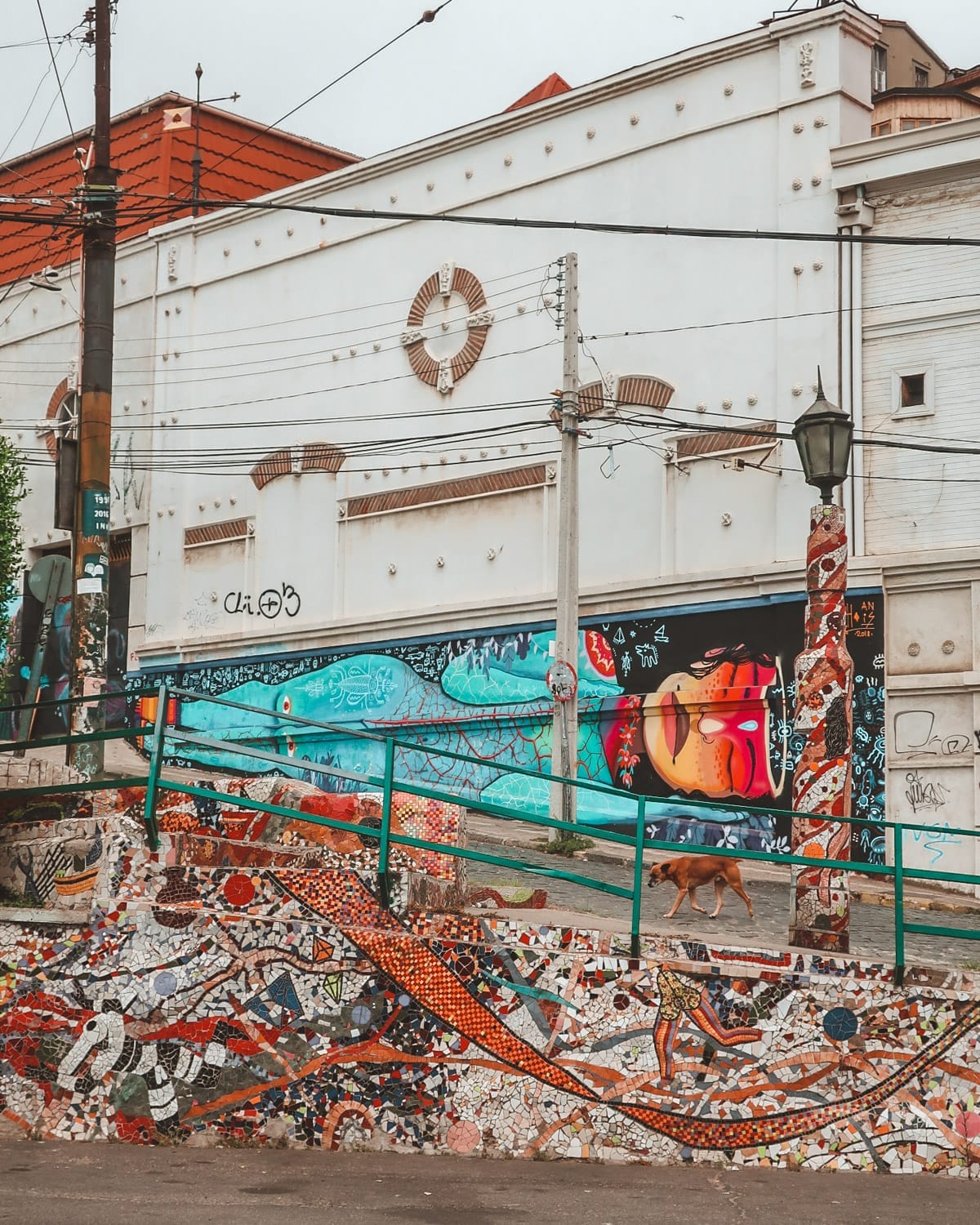 Street art in Valparaíso featuring a blue and orange mural on a white building, mosaic steps, and a dog walking uphill.