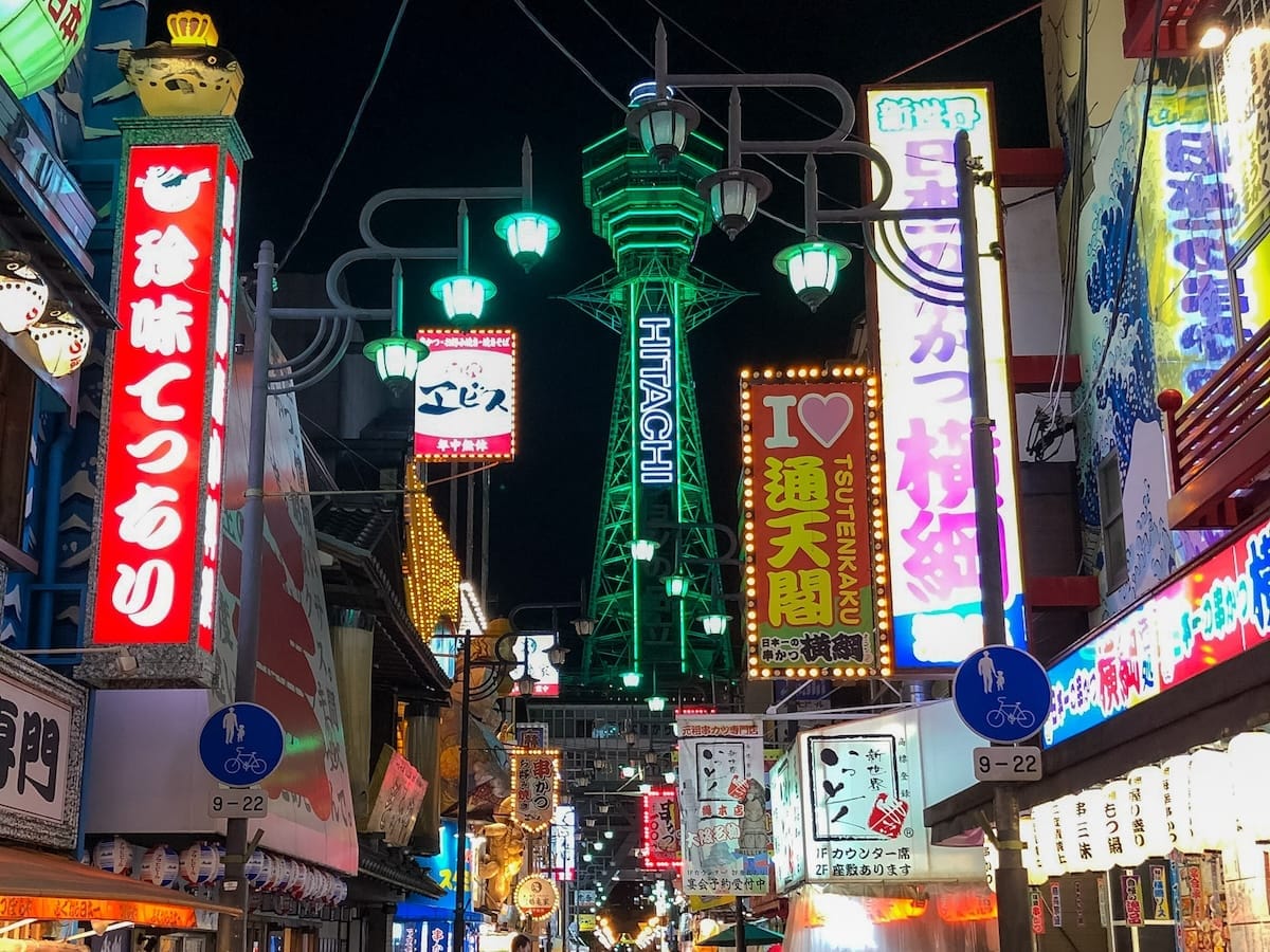 A vibrant Osaka street scene at night with neon signs in Japanese and Tsutenkaku Tower lit up in green in the background.
