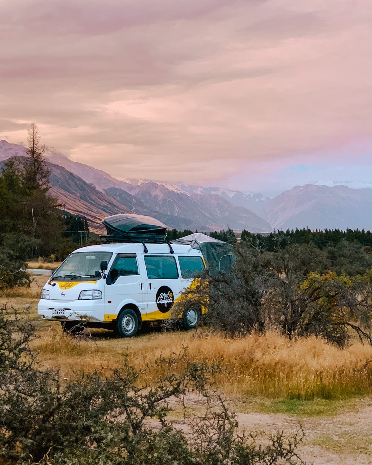 Campervan with rooftop gear parked in golden grass on New Zealand’s South Island, with purple mountains lit up from the sunset.