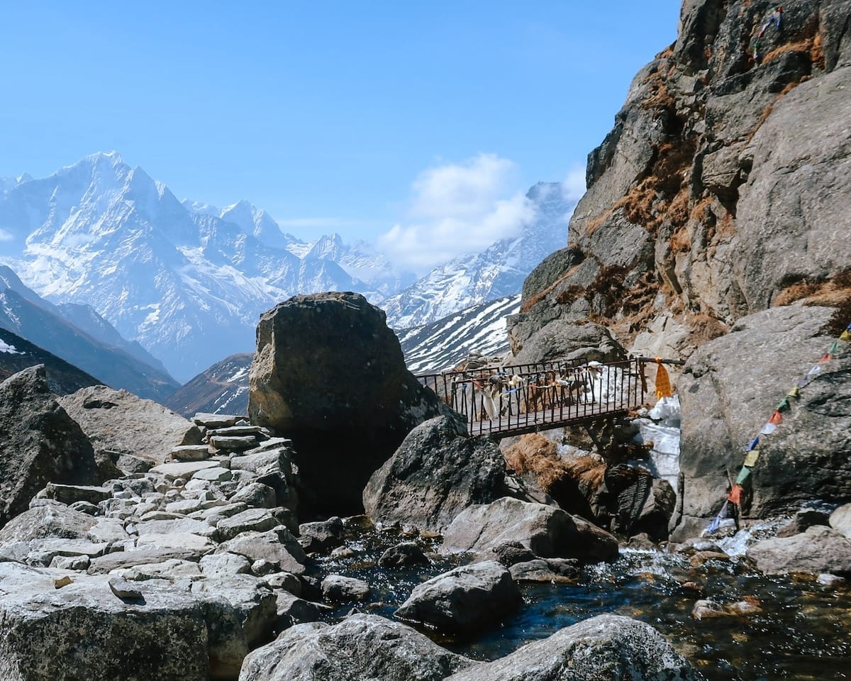Small suspension bridge over a stream, surrounded by boulders, with towering peaks in the distance.