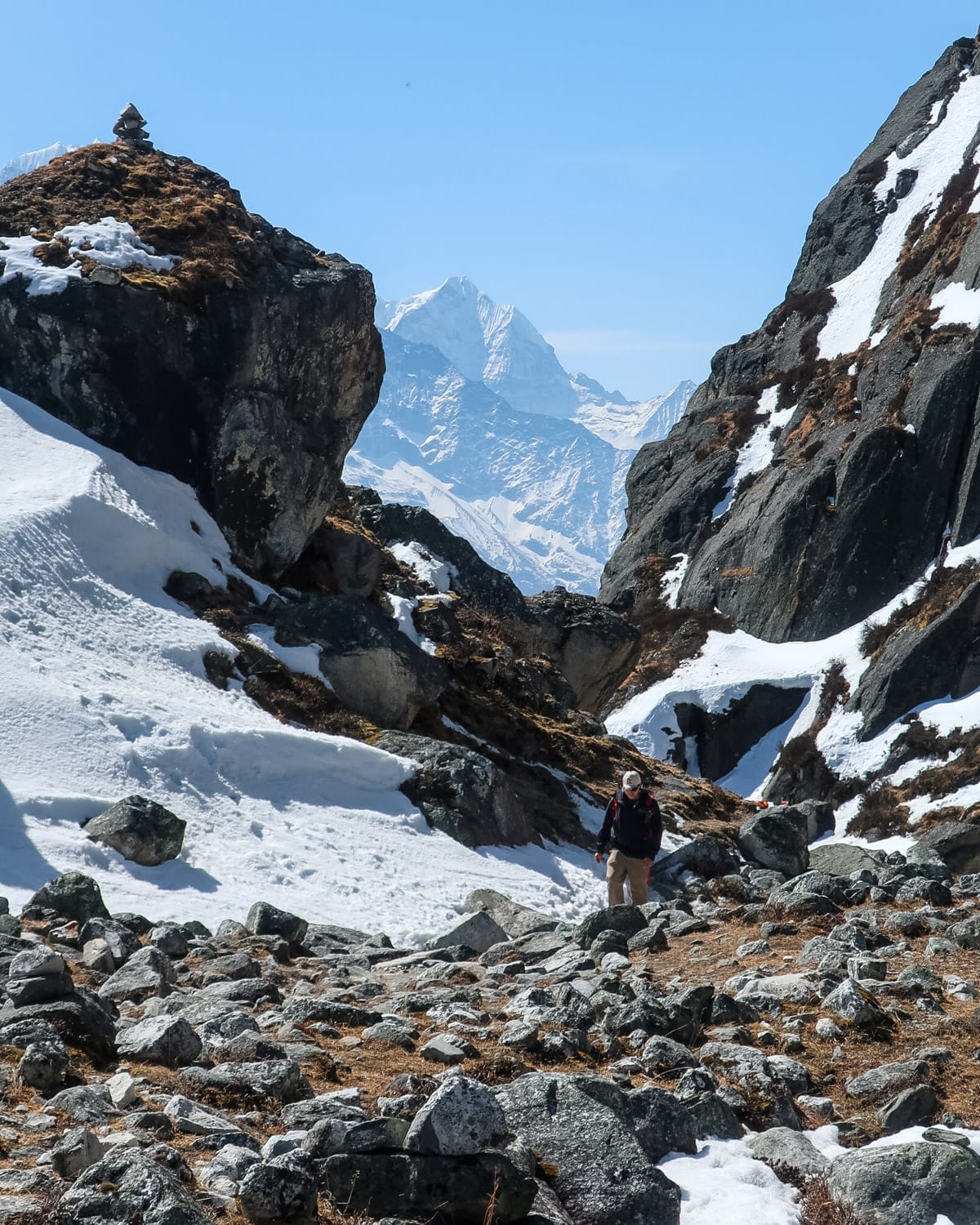 Trekker walks among boulders through a snowy mountain pass with sharp Himalayan peaks rising behind.