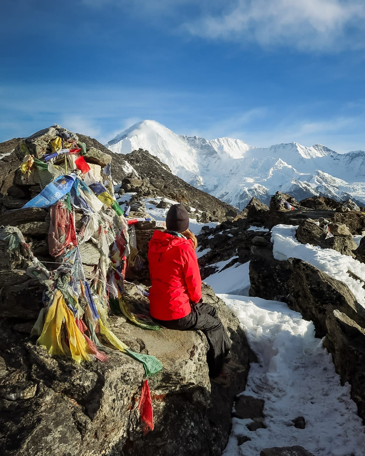 Cec in red jacket sits beside colourful prayer flags at the summit of Gokyo Ri, gazing at snowy peaks under a blue sky.