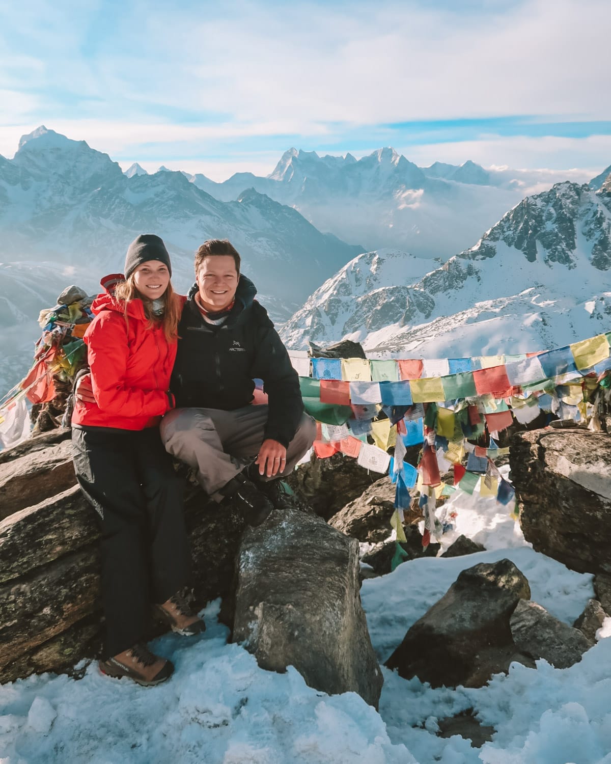 Ari and Cec sitting on rocks by prayer flags atop Gokyo Ri with a panoramic view of snowy Himalayan peaks.