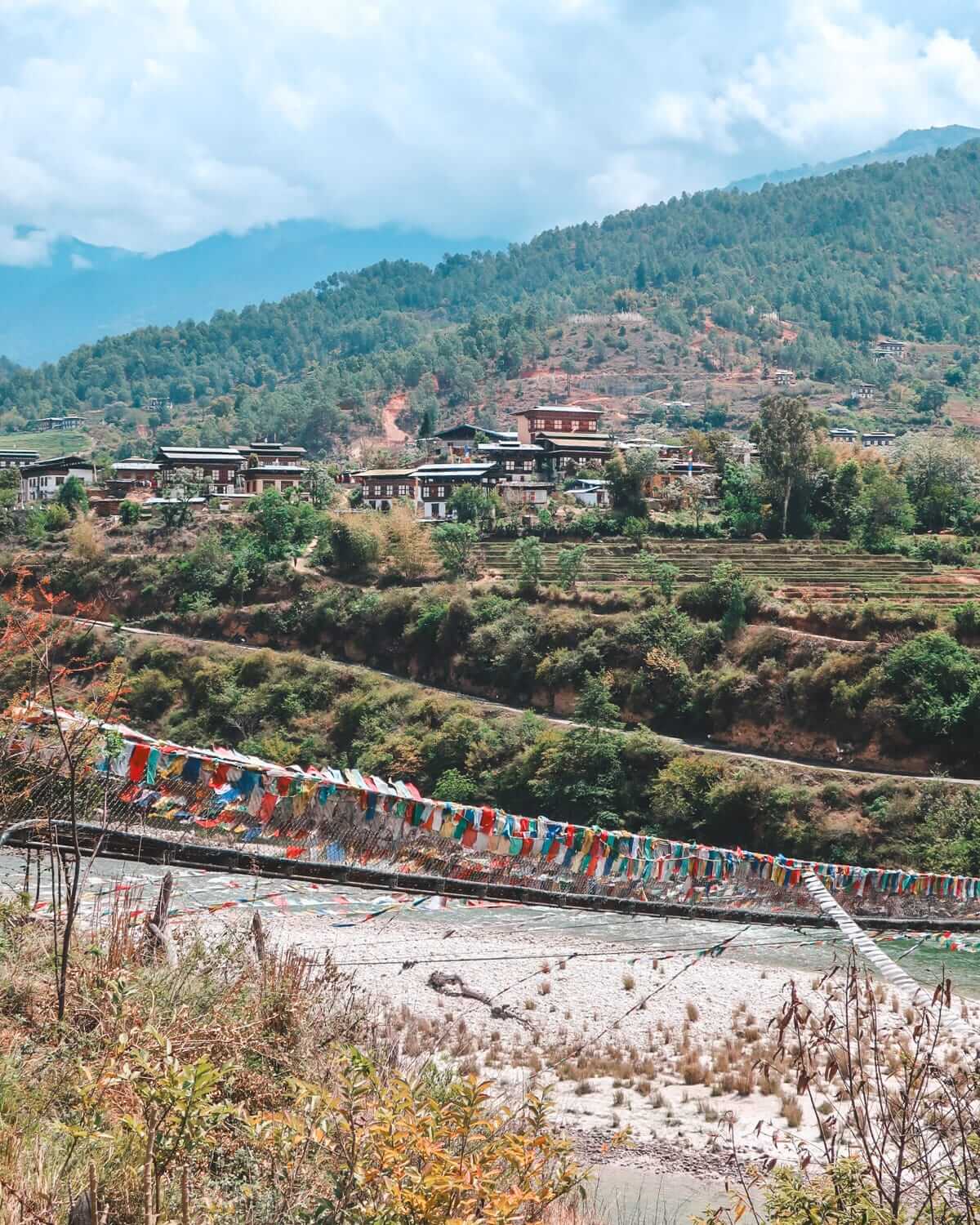 Suspension bridge in Bhutan covered in prayer flags, stretching across a river with a hillside village and lush greenery in the distance.