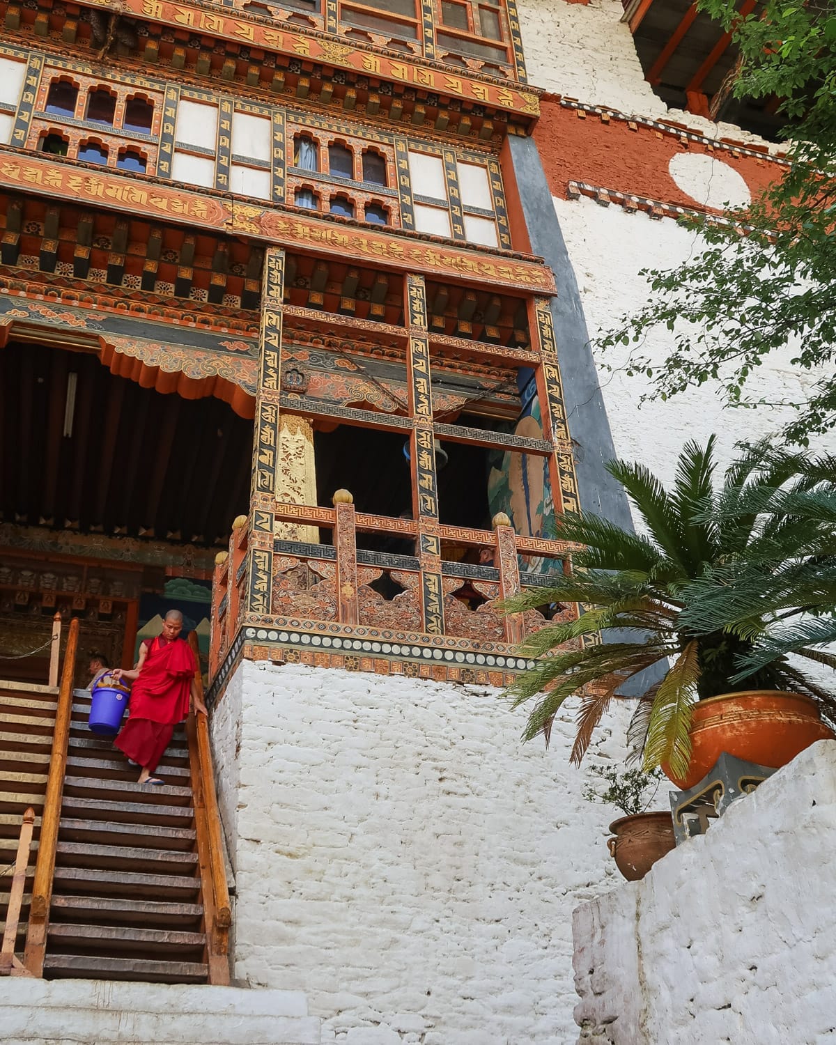Monk in a red robe descending temple stairs, carrying a blue bucket, with a backdrop of Bhutanese architecture and decorative details.