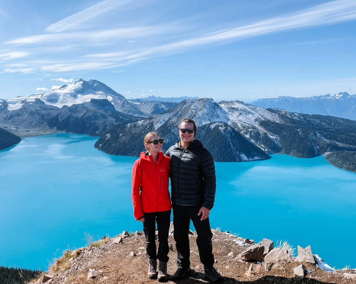 Cec and Ari standing on a rocky peak overlooking the bright blue Garibaldi Lake, surrounded by snow-capped mountains.