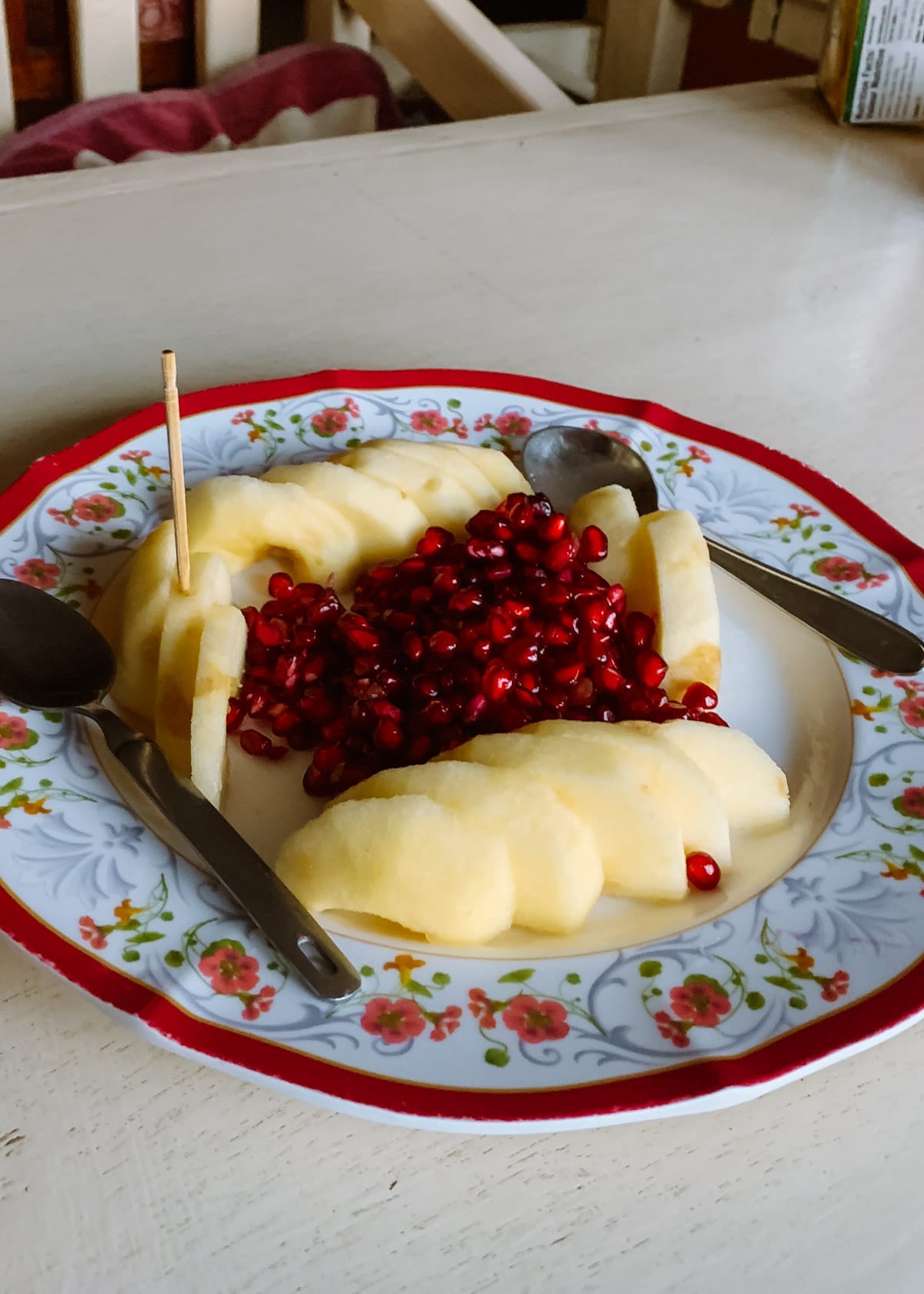 Sliced apples and pomegranate seeds served on a floral plate with two spoons and a toothpick.