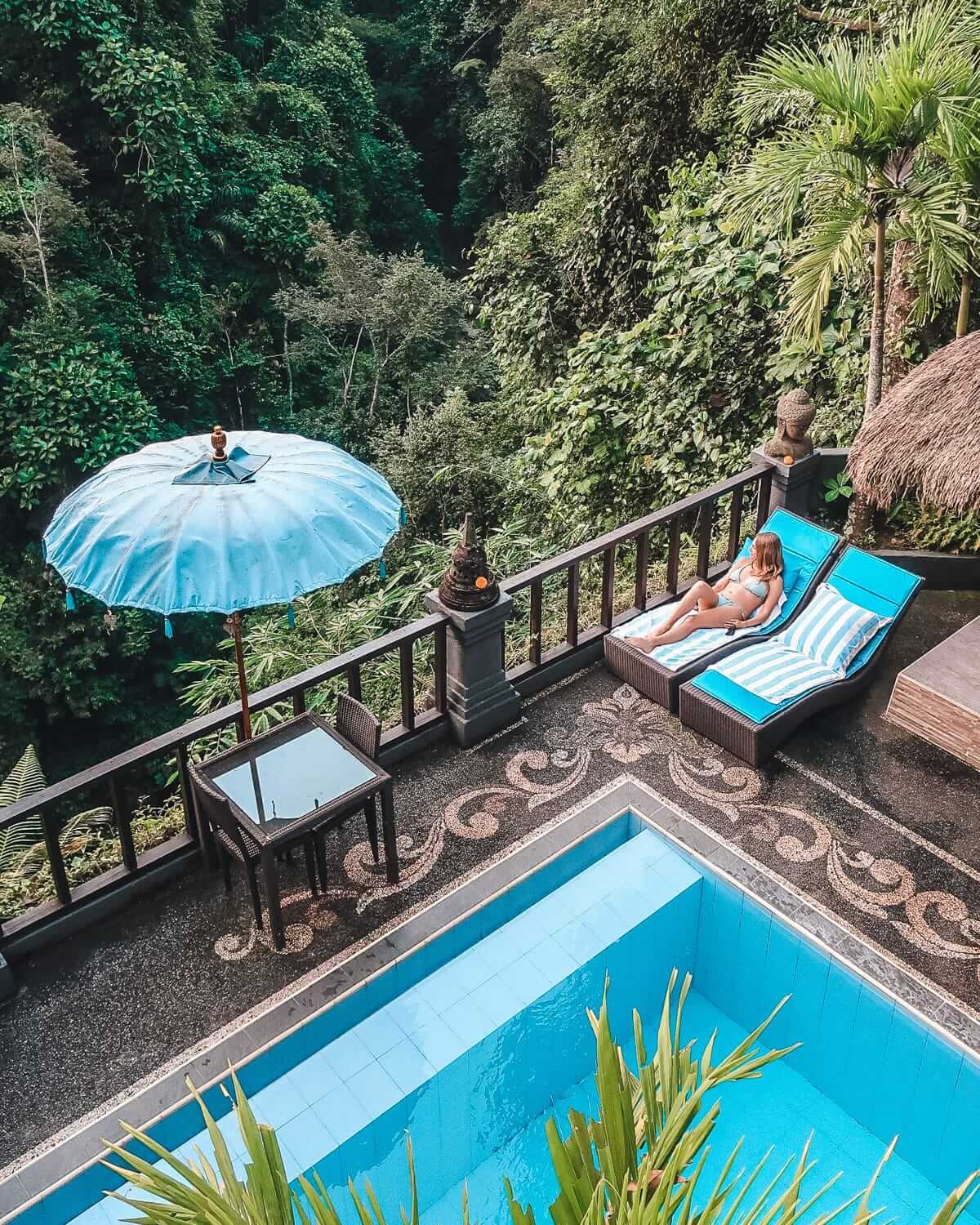 Cec lounges on a sunbed beside a pool, overlooking a lush tropical jungle with a blue umbrella in Ubud.