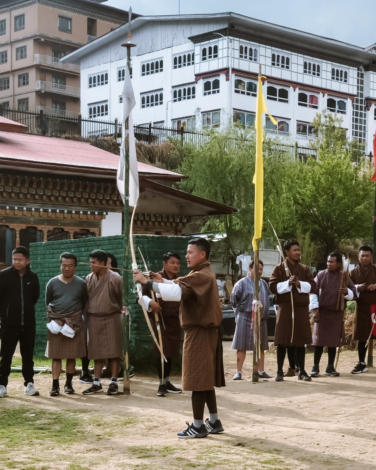 Bhutanese archery competition, where men in traditional gho attire gather as a participant aims with a bow in an open field.