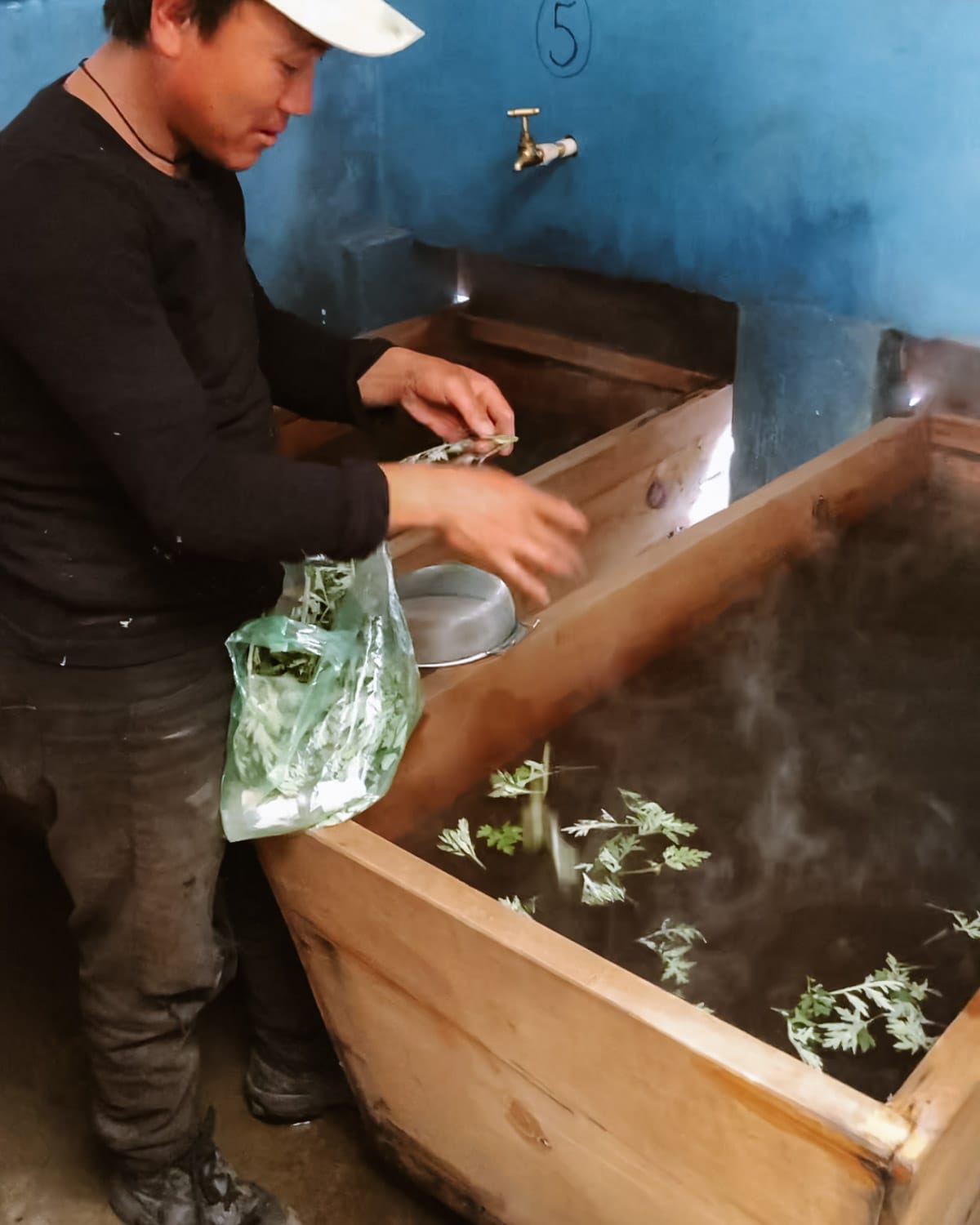 Bhutanese man preparing a herbal hot stone bath, adding artemisia leaves to steaming water inside a wooden tub.