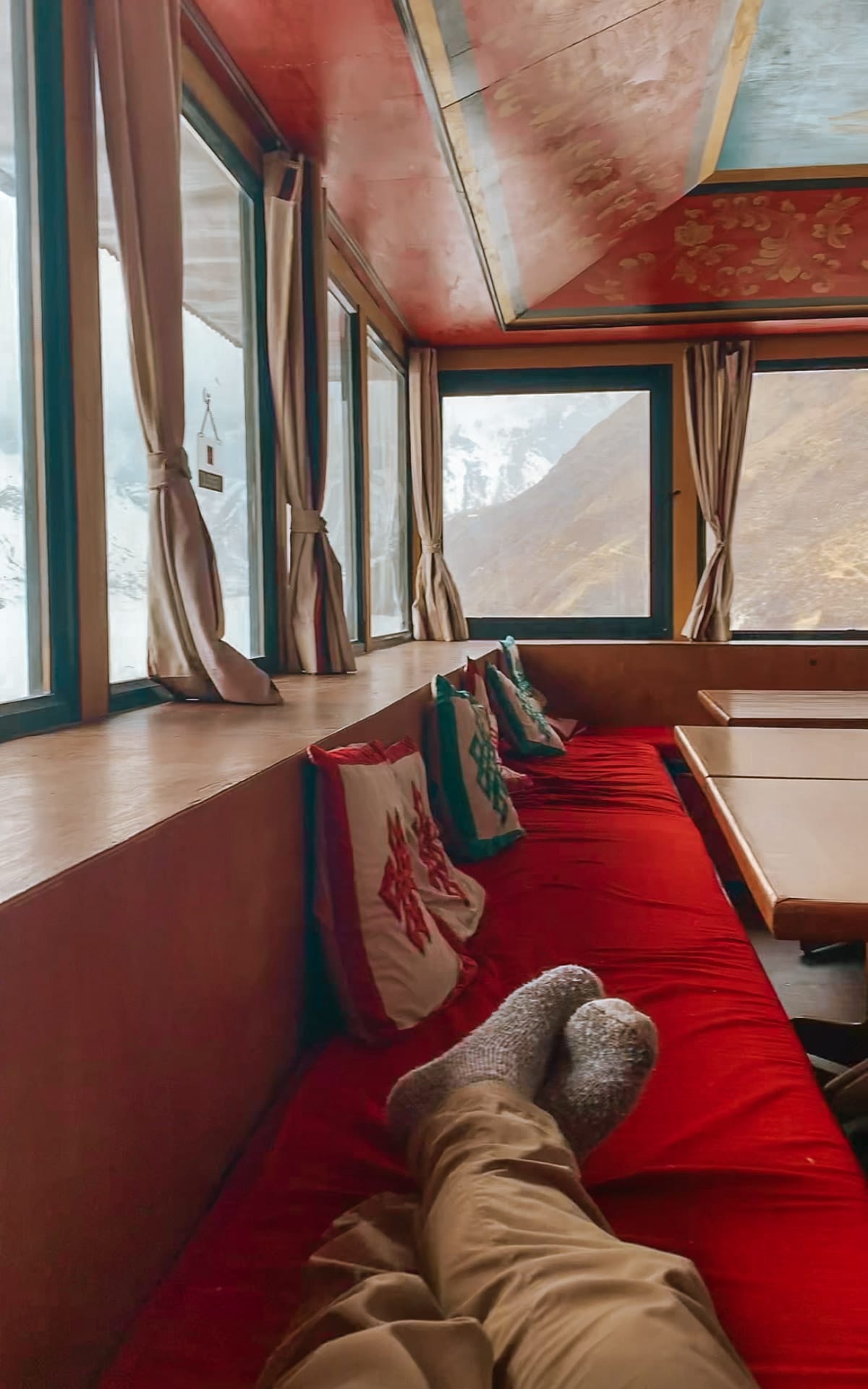 View from a cozy teahouse bench in Gokyo with red cushions and pillows, overlooking snowy mountains through large windows.