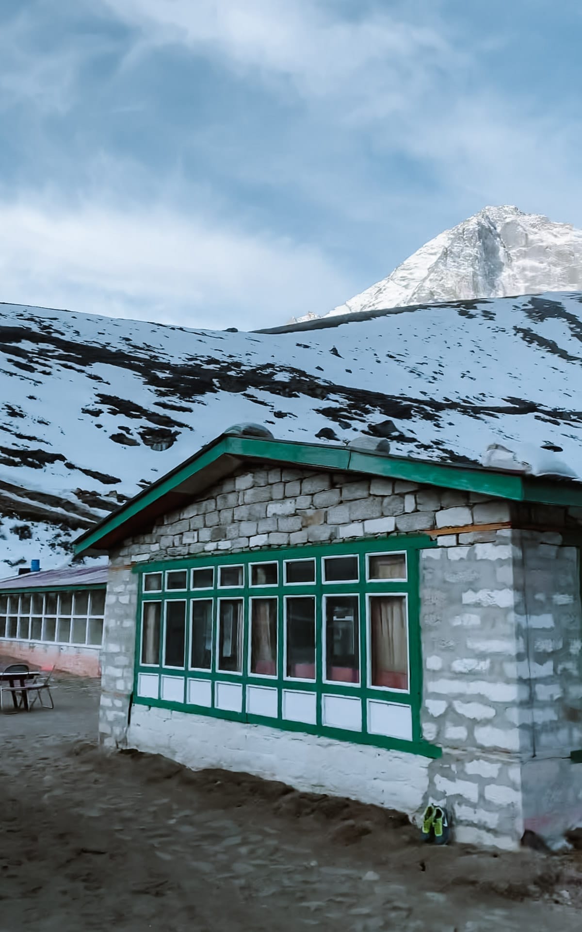 Stone teahouse with green trim in the village of Machermo stands at the base of a snowy slope with a Himalayan peak in the background.