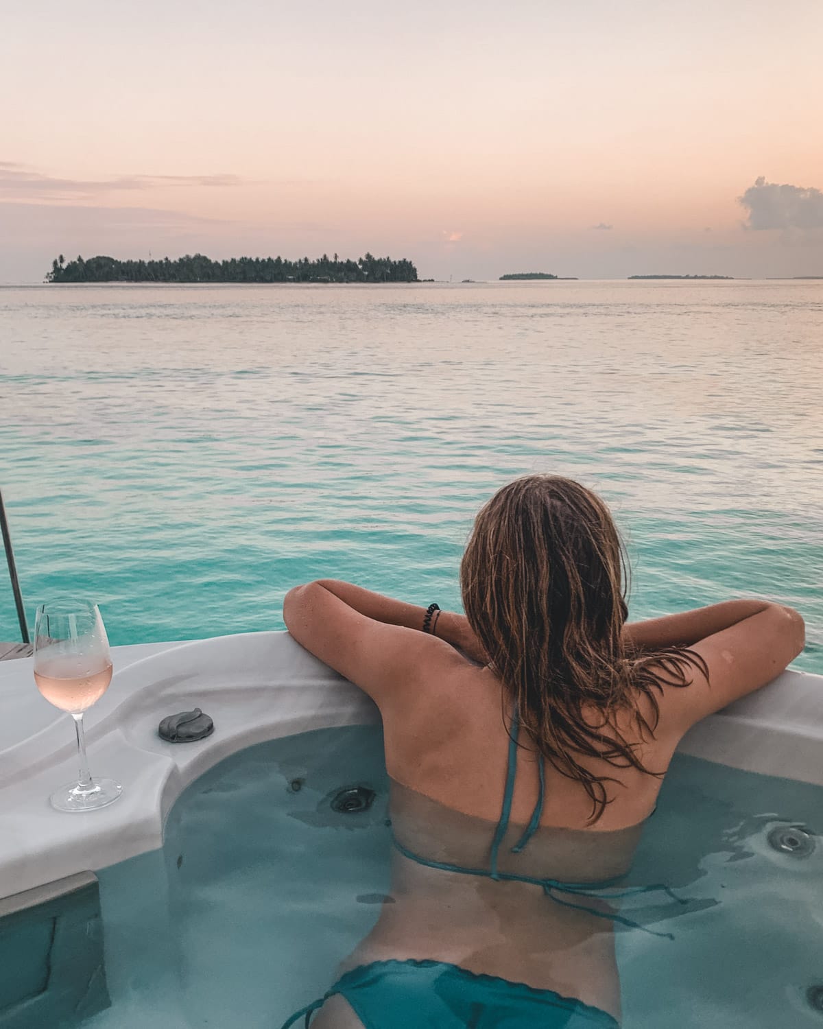 Cec soaking in a private jacuzzi at sunset with a glass of rose, overlooking calm ocean waters and a distant island.